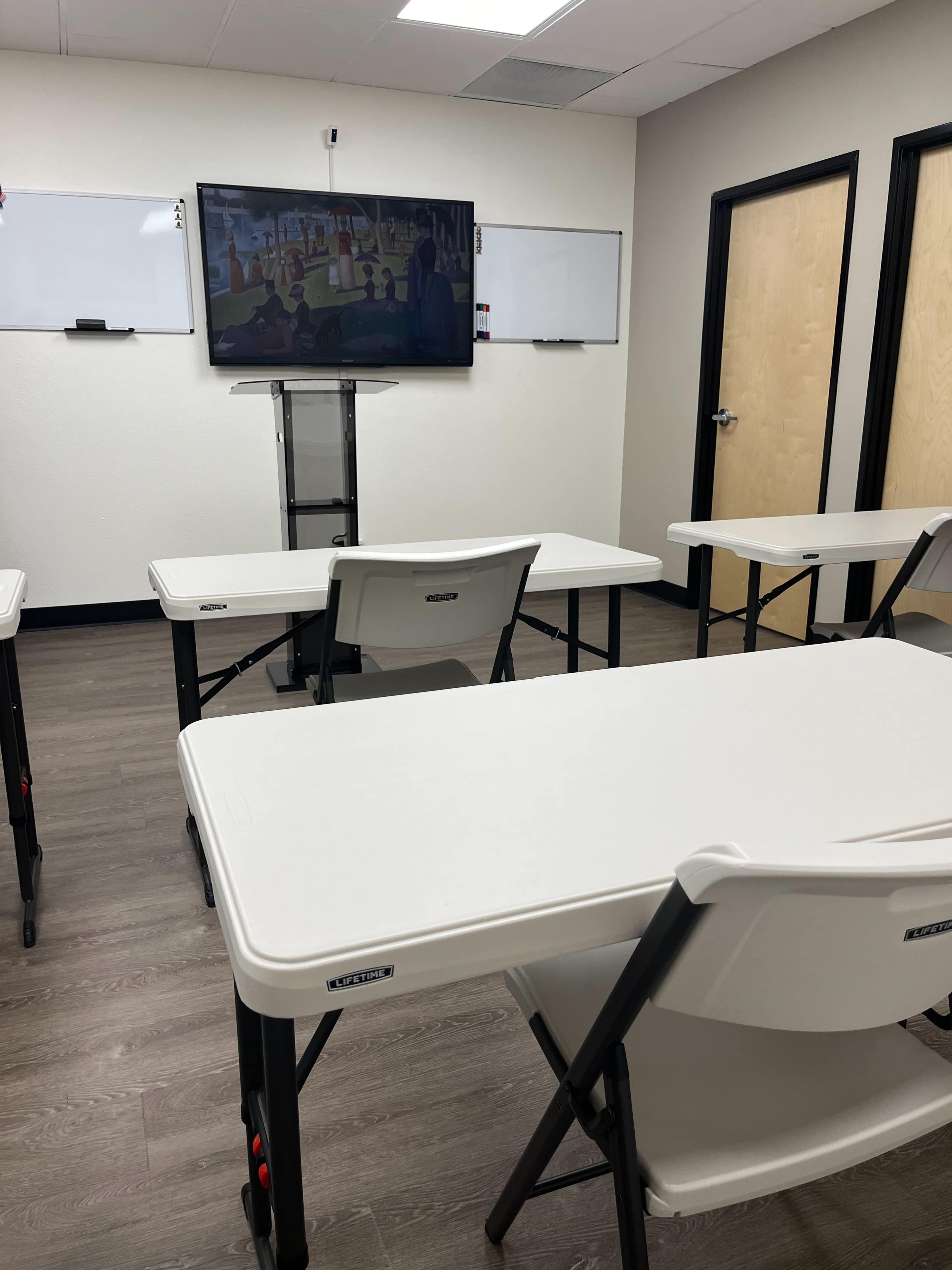 A classroom with several white folding tables arranged for use, a large screen displaying an image at the front, and whiteboards on the walls.