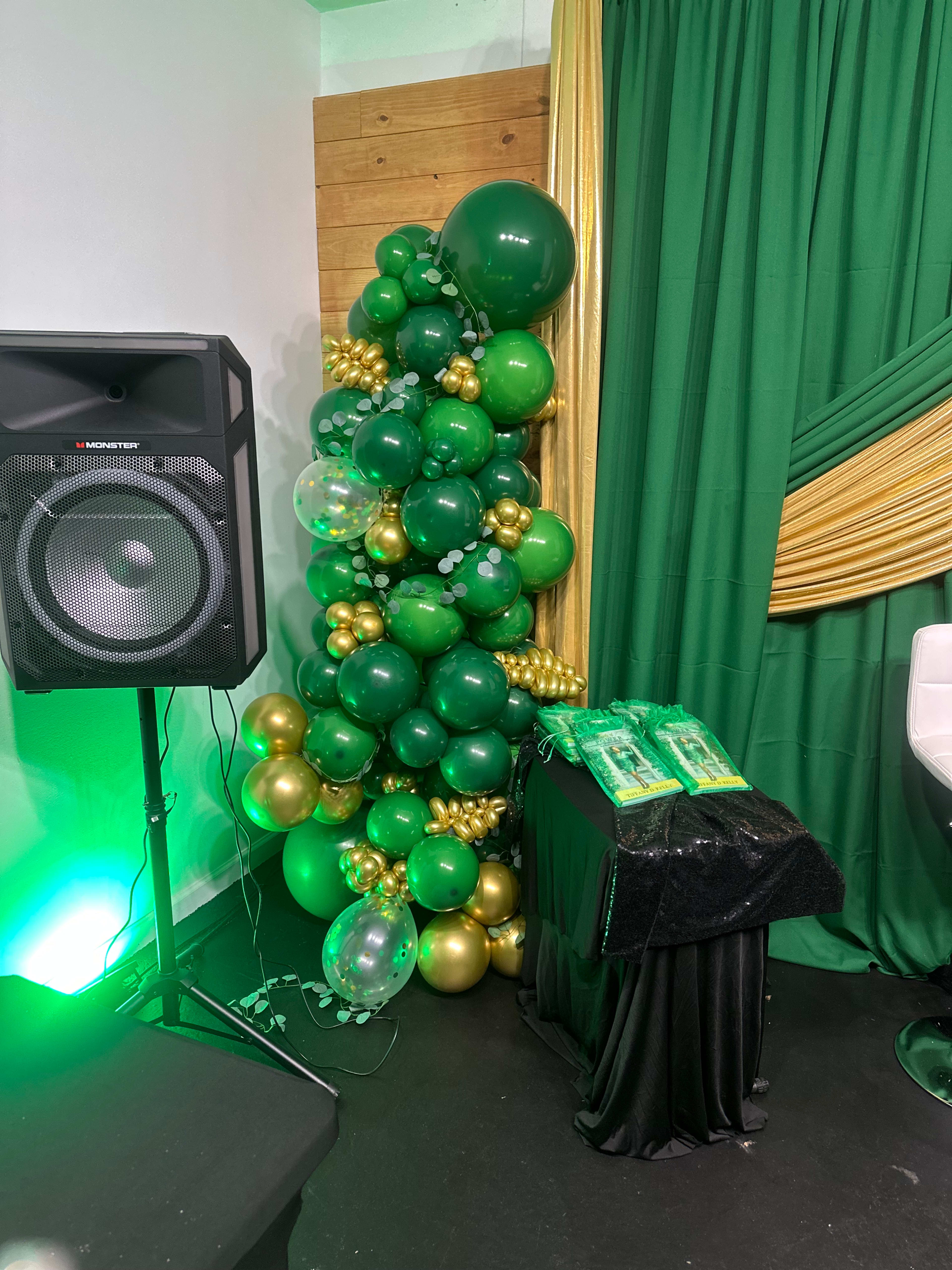 The image shows a decorative arrangement of green and gold balloons next to a speaker and a table covered with a black cloth.
