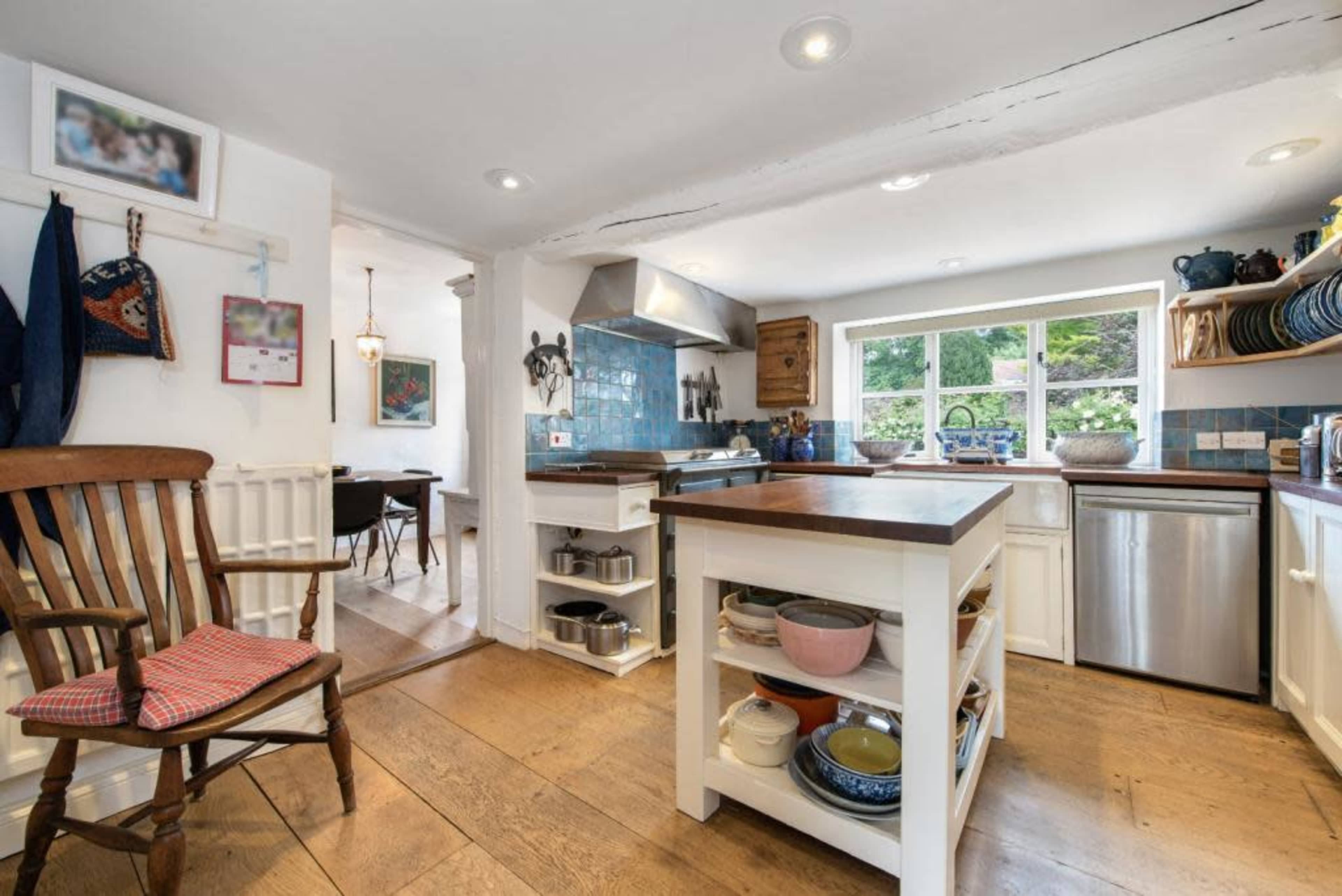 A spacious kitchen features a central island with various cooking utensils, wooden flooring, and a large window overlooking an outdoor area.
