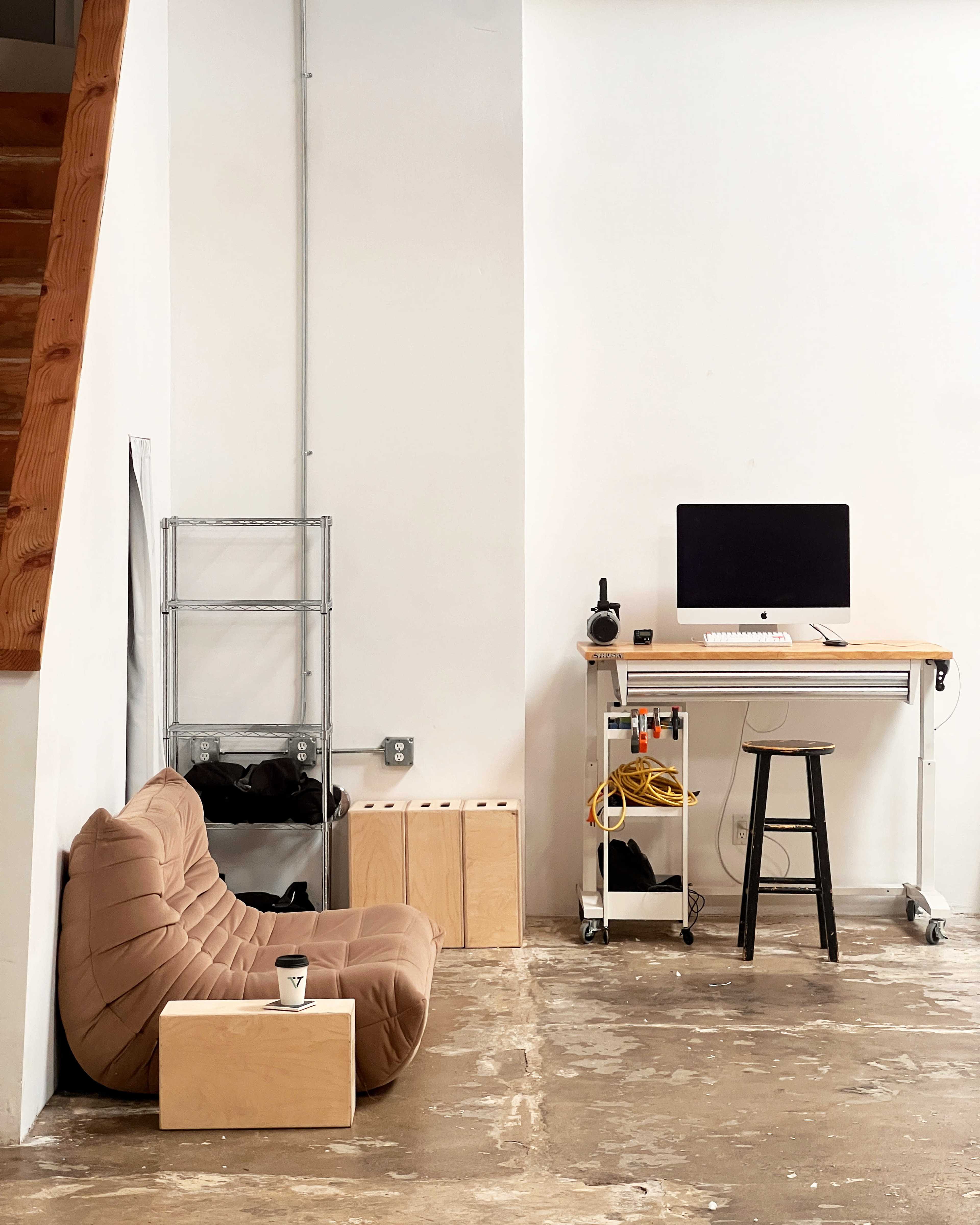 A minimalistic workspace features a wooden desk with a computer, a black stool, a cushioned seating area, and storage boxes against a white wall.