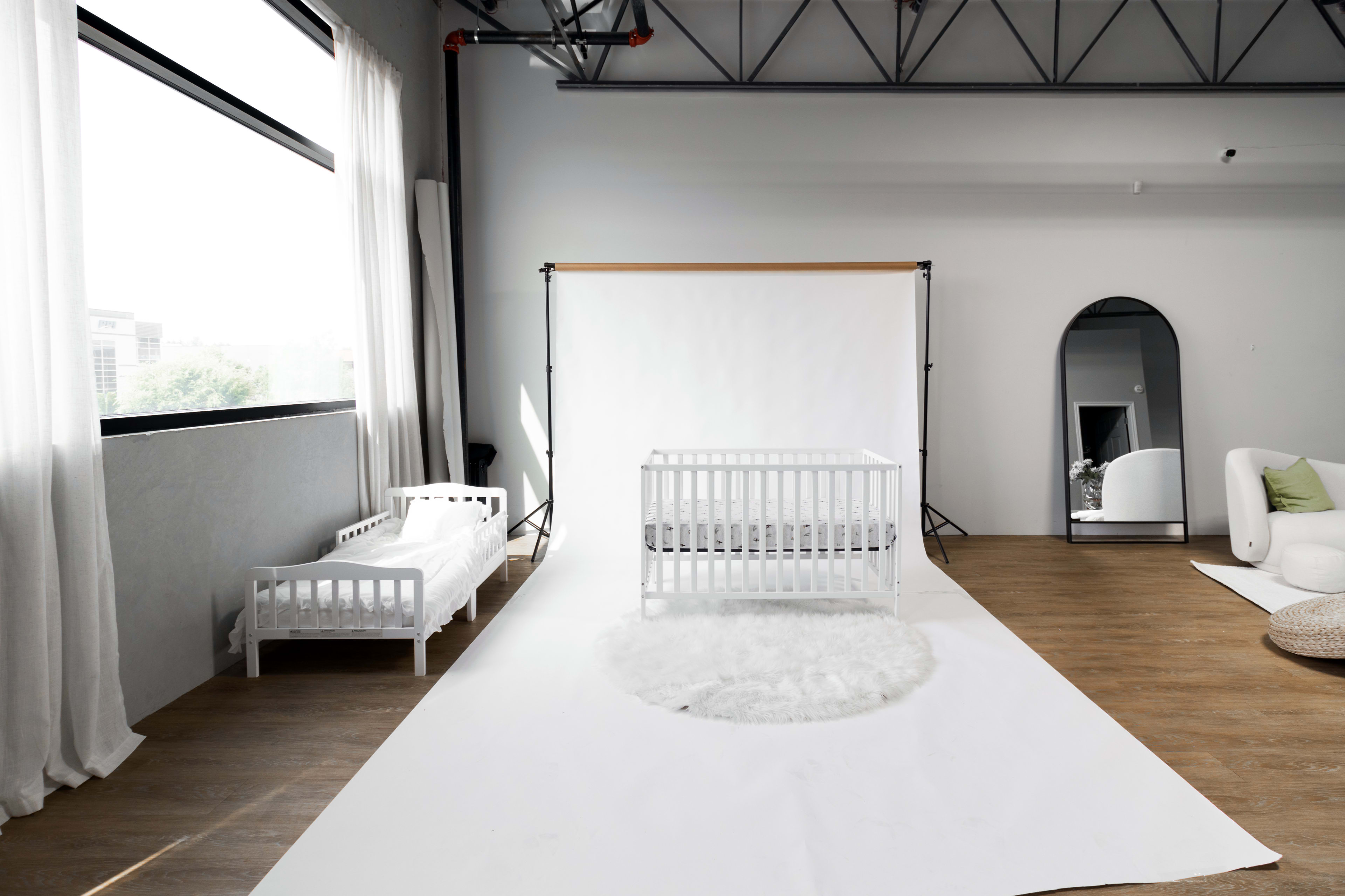 A bright, minimalist room features two white cribs, a round rug, and a large mirror against a light-colored backdrop.