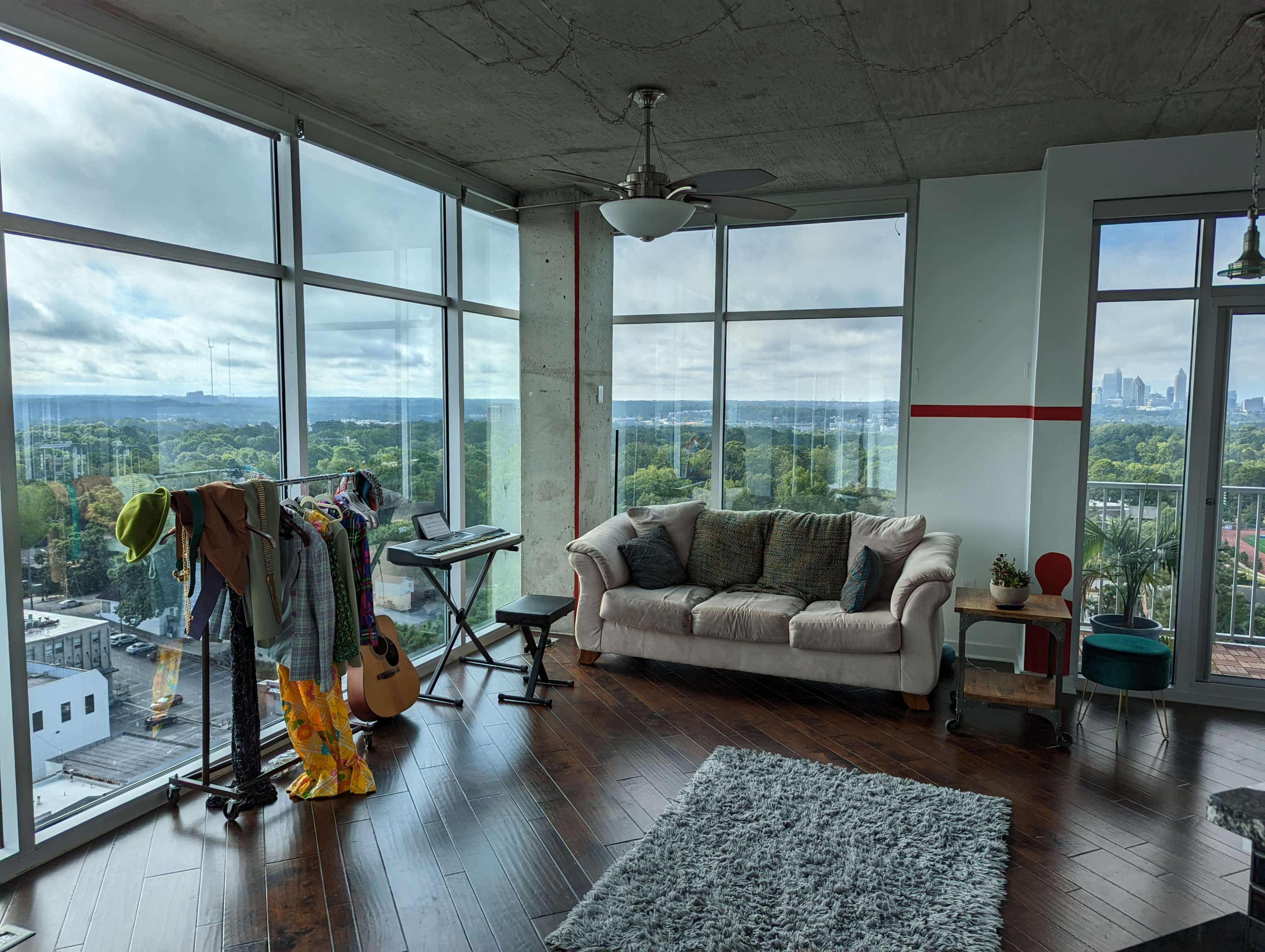 A modern living room with a couch, a clothing rack, a guitar, and large windows offering a view of a skyline and landscape beyond.