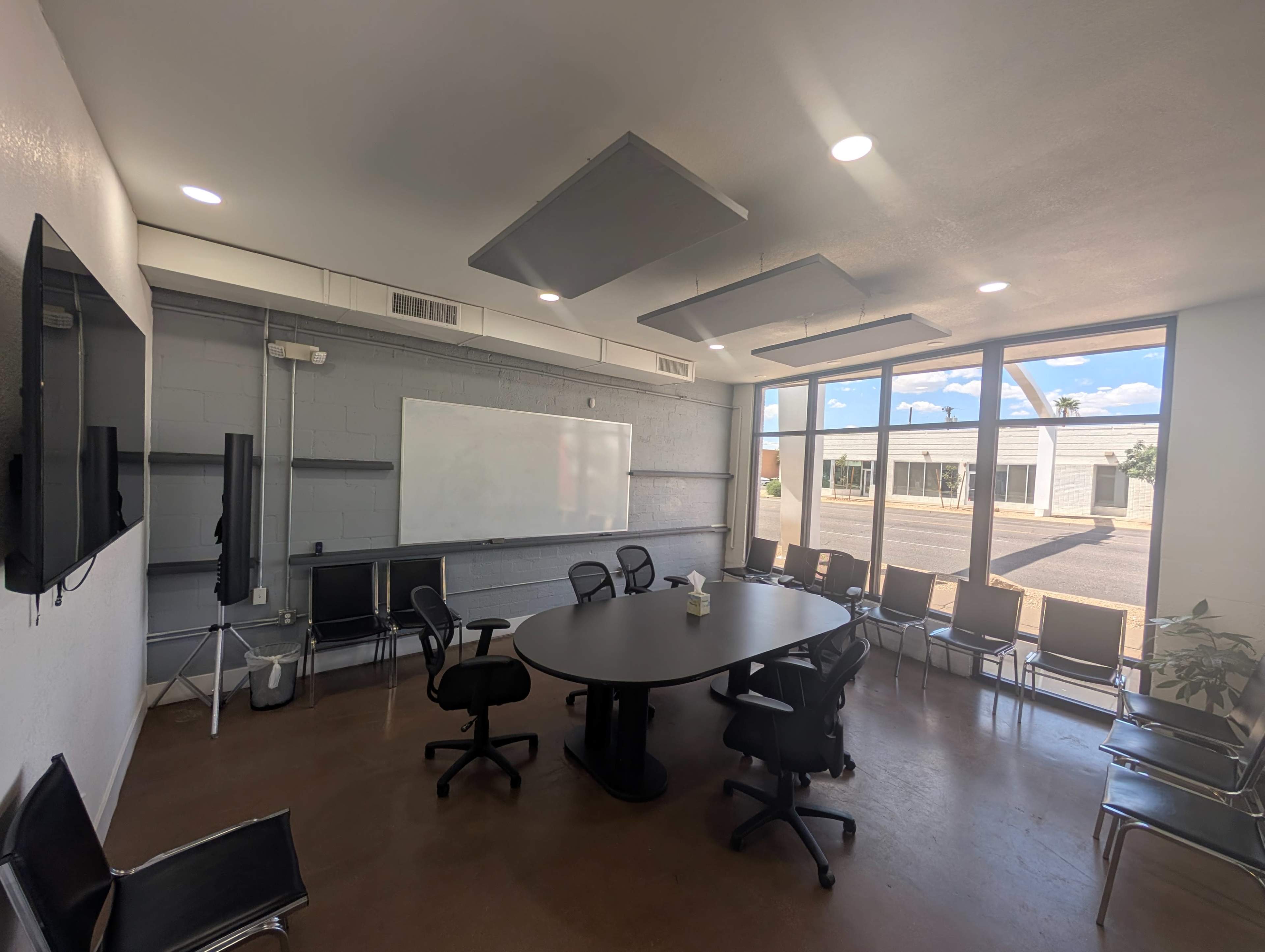 A conference room features a large round table surrounded by chairs, a whiteboard, and two wall-mounted TVs, with windows providing natural light and a view of the exterior.