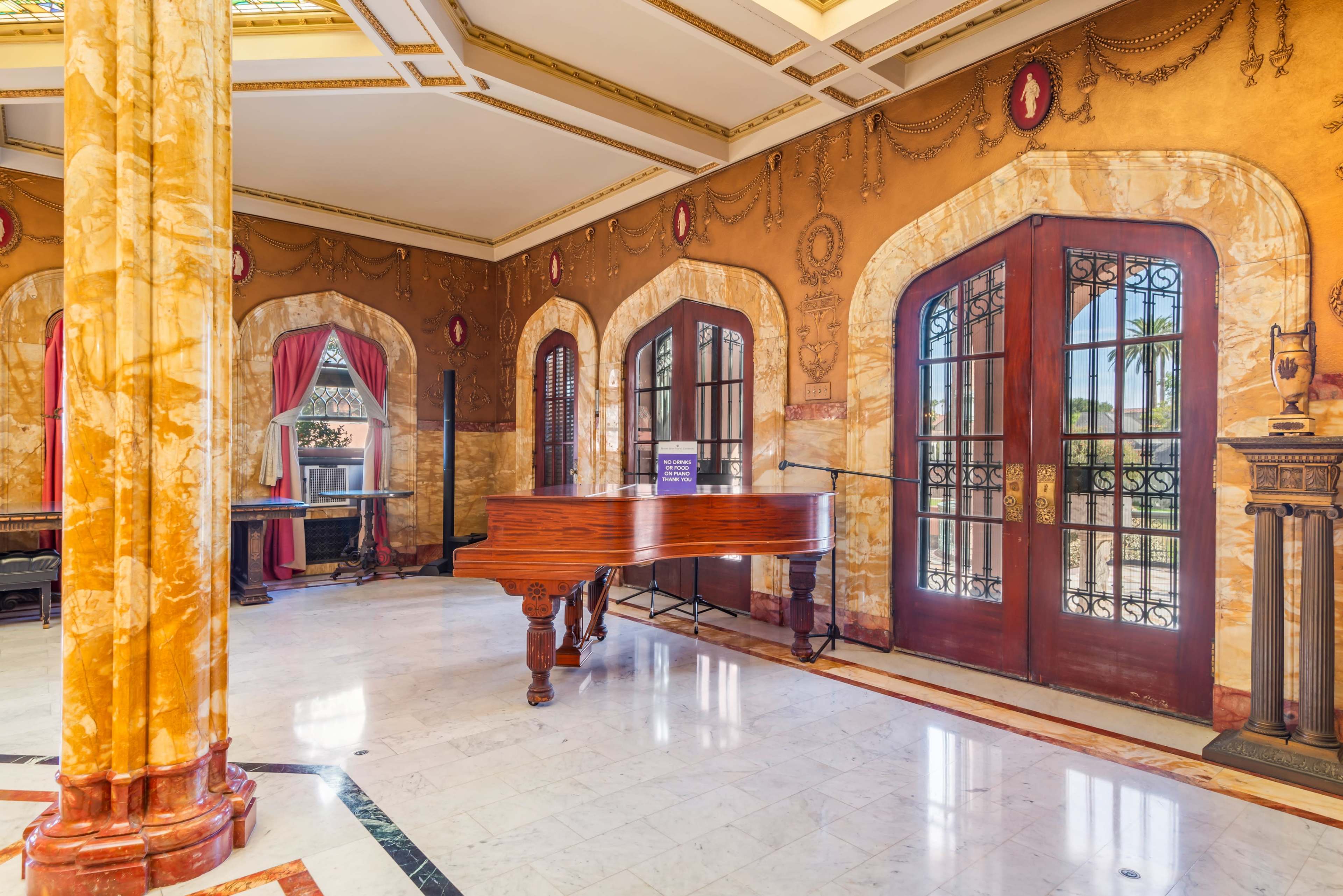 The image shows a grand room featuring a polished wooden piano, marble flooring, and large arched windows framed by ornate curtains.