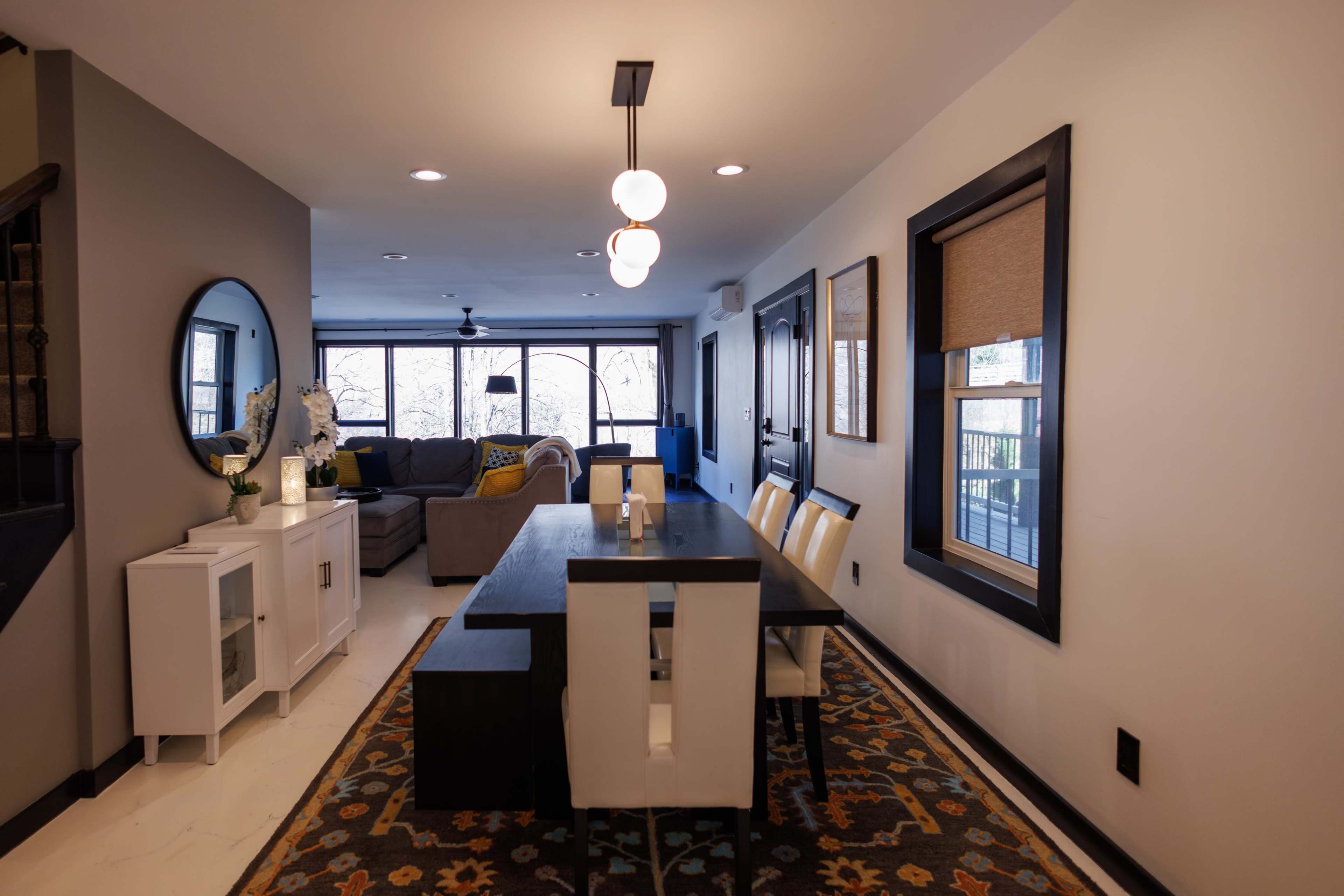A dining area features a long table surrounded by white chairs, adjacent to a living space with a gray sofa and large windows.