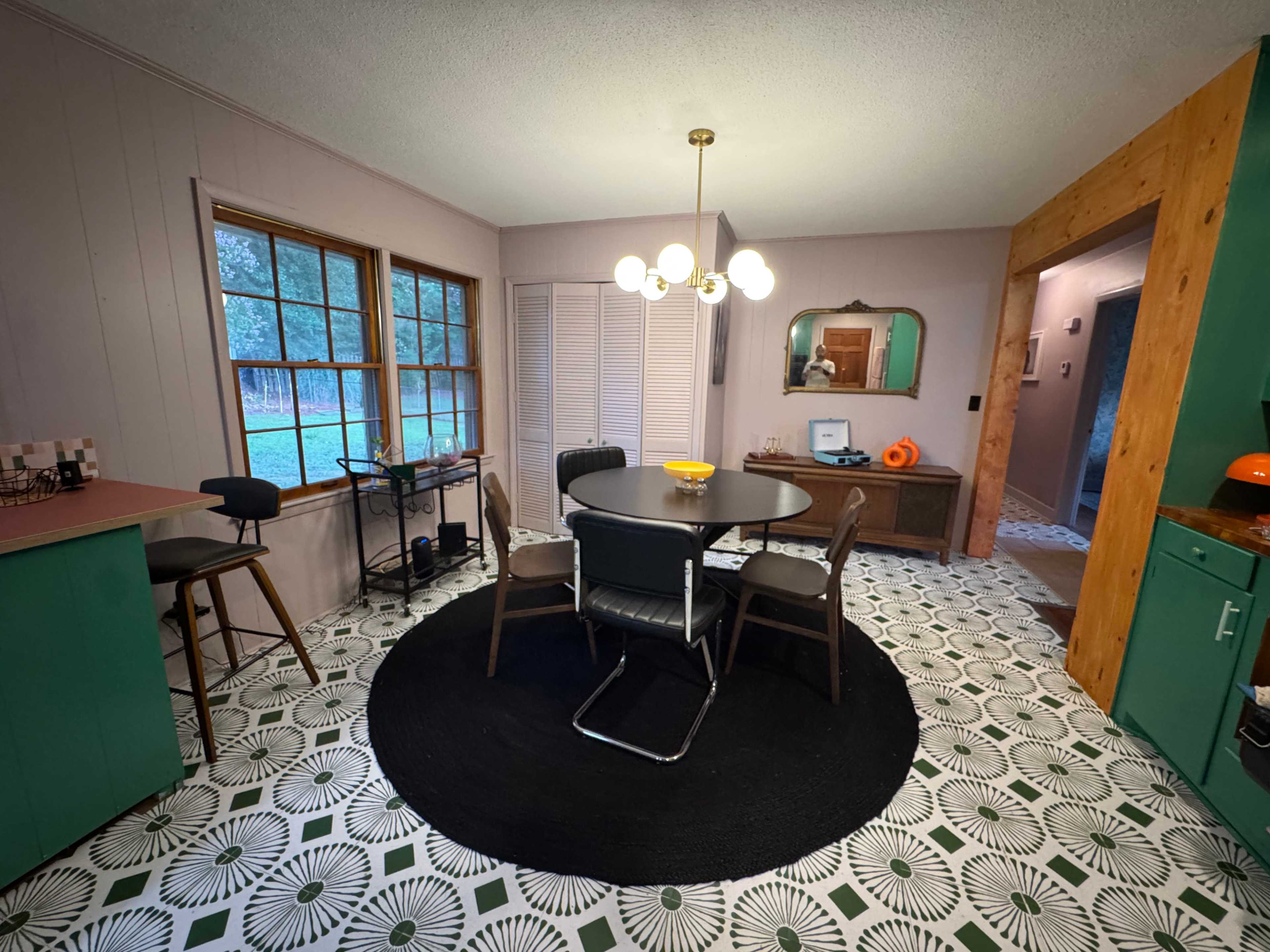 A modern kitchen with a circular dining table surrounded by four chairs, featuring large windows and a patterned floor.
