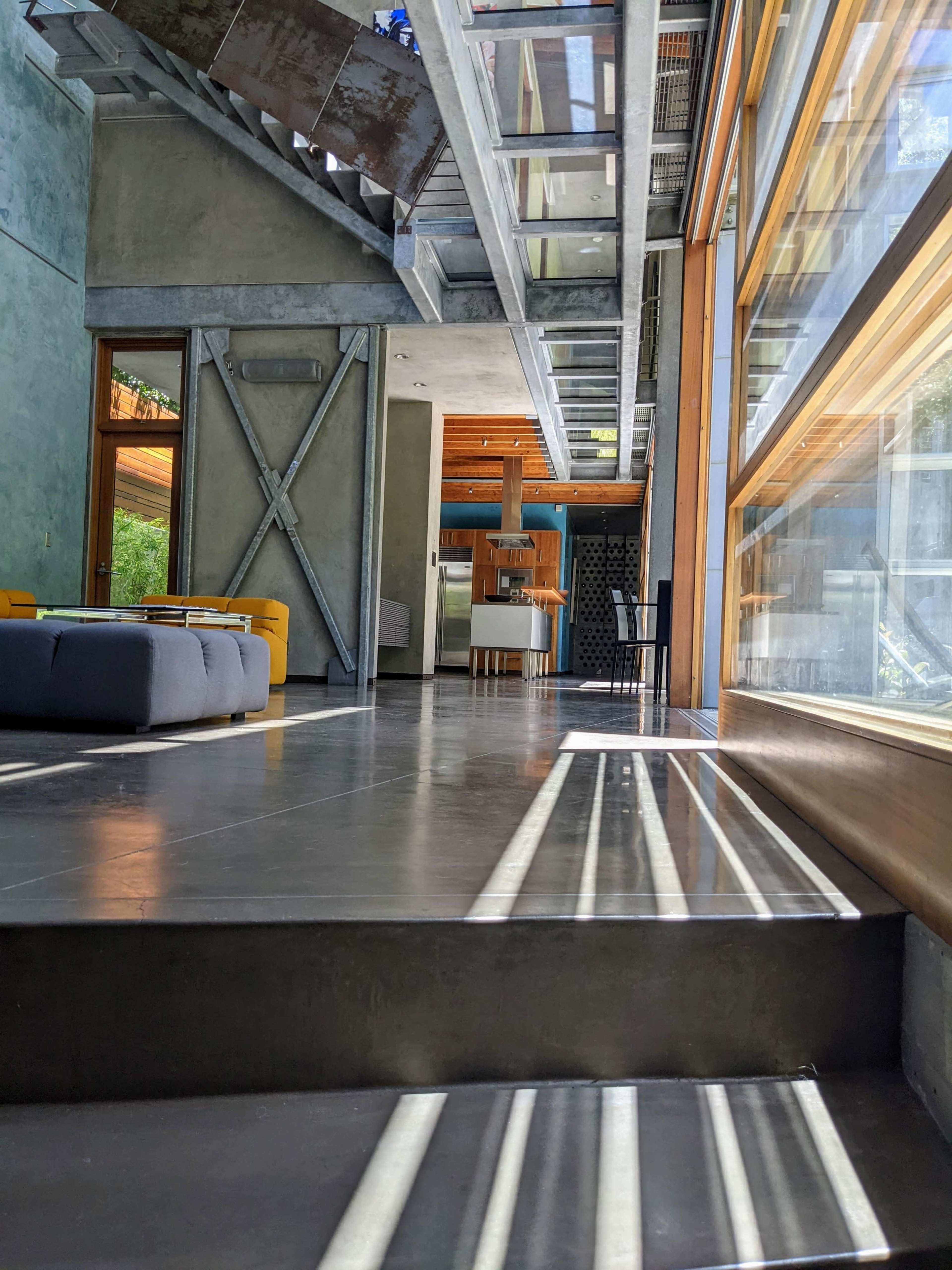 The image shows a modern interior space featuring concrete walls, large windows allowing natural light, and a staircase leading to a lower level.