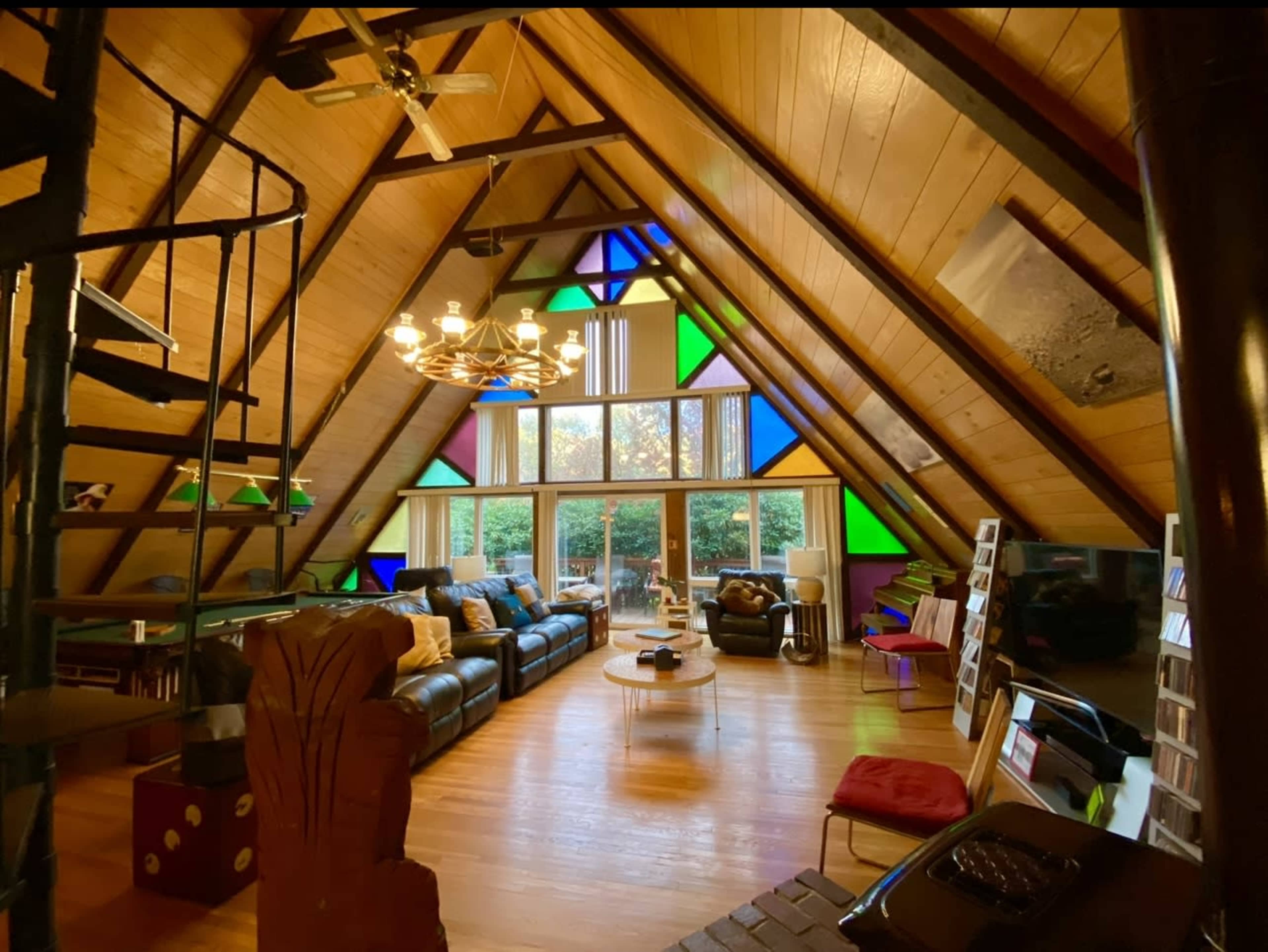 The interior of a spacious, triangular room features a wooden ceiling with large windows adorned with colorful stained glass, modern furniture, and a spiral staircase.