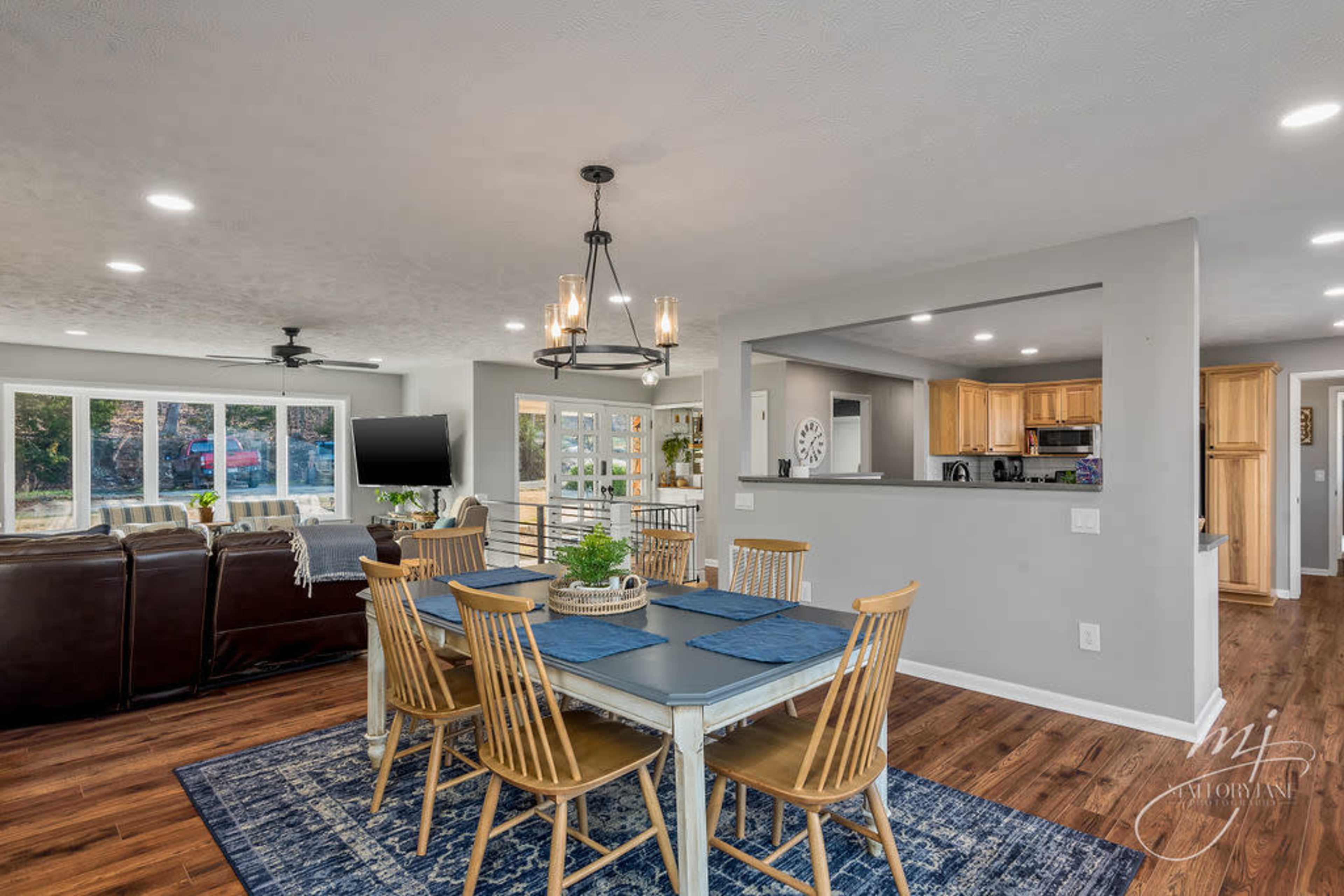 The image shows a modern dining area with a wooden table and chairs, a large area rug, and an adjoining kitchen visible in the background.