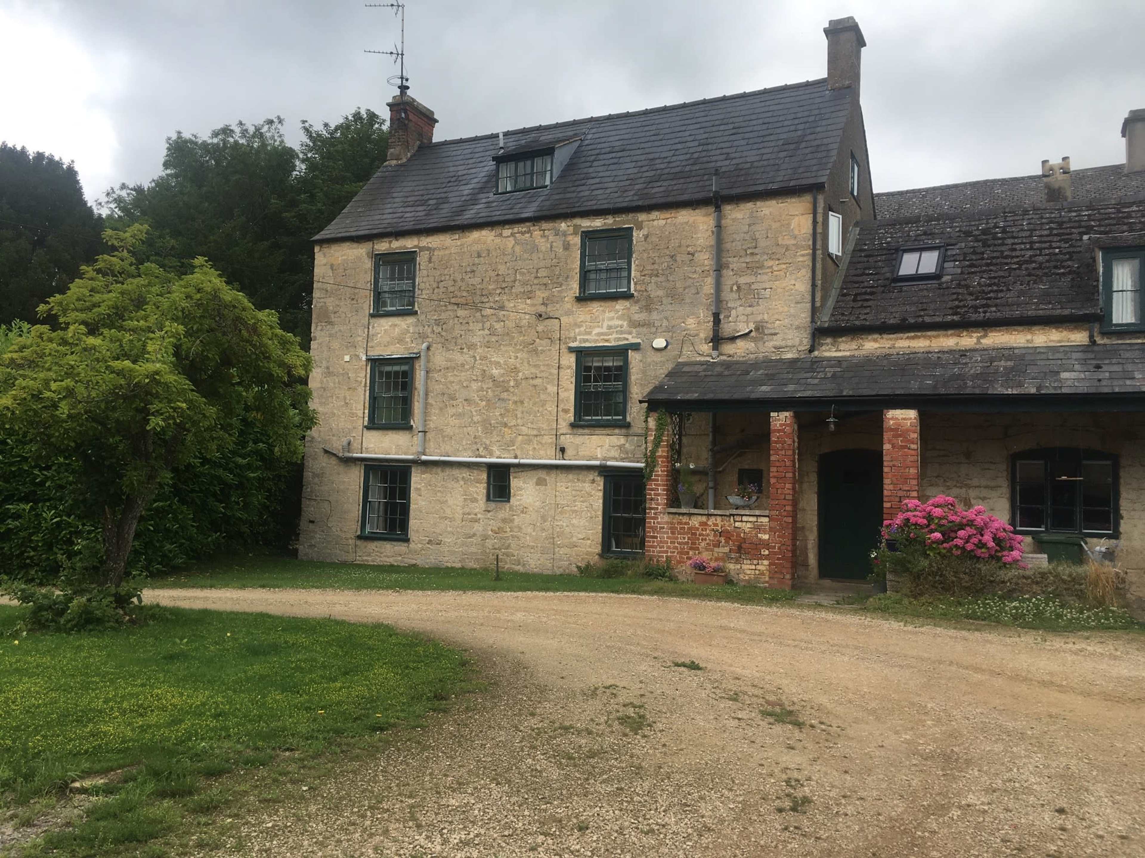 A stone house with multiple windows and a gravel driveway is set against a backdrop of greenery.