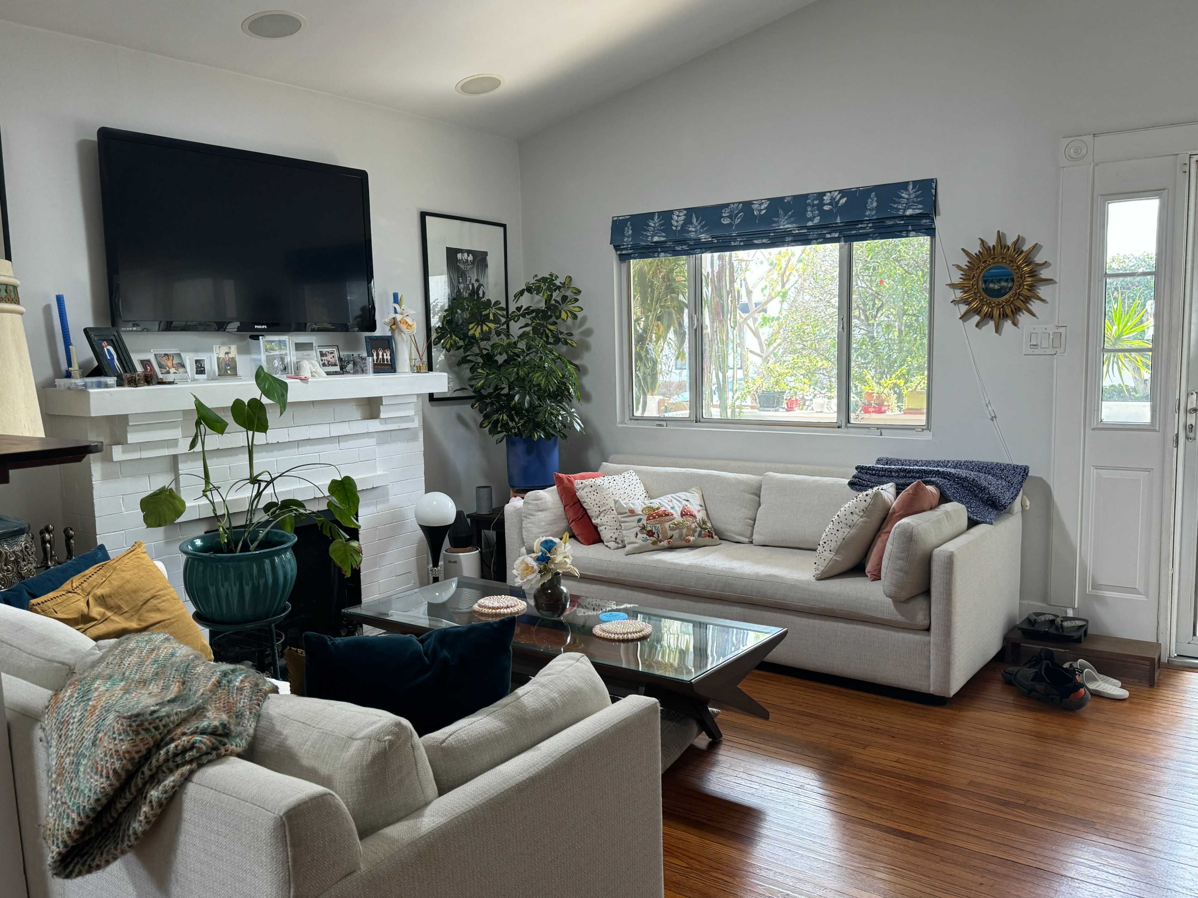 The image shows a living room with two light-colored sofas, a coffee table, a television, and large windows allowing natural light, surrounded by indoor plants and decorative items.