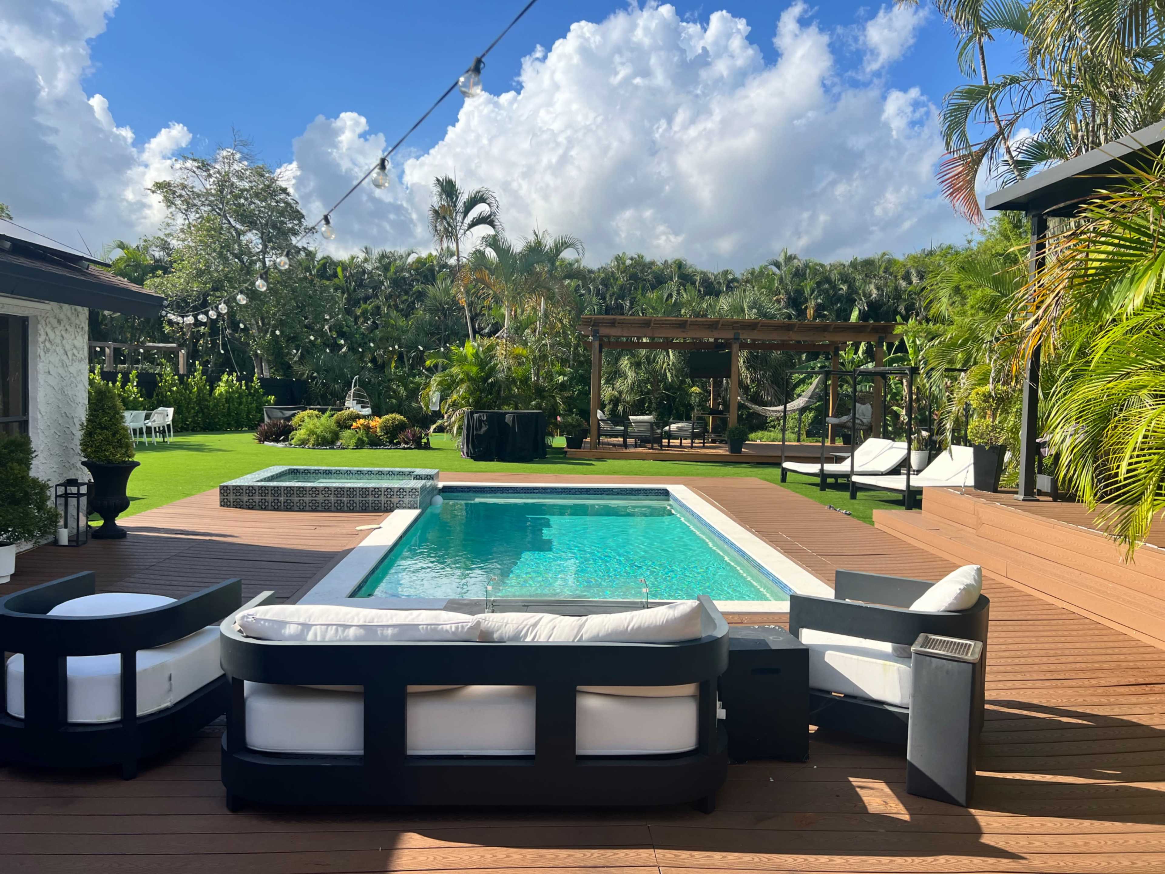 The image shows a sunny backyard with a pool surrounded by wooden decking, lounge chairs, and lush greenery.