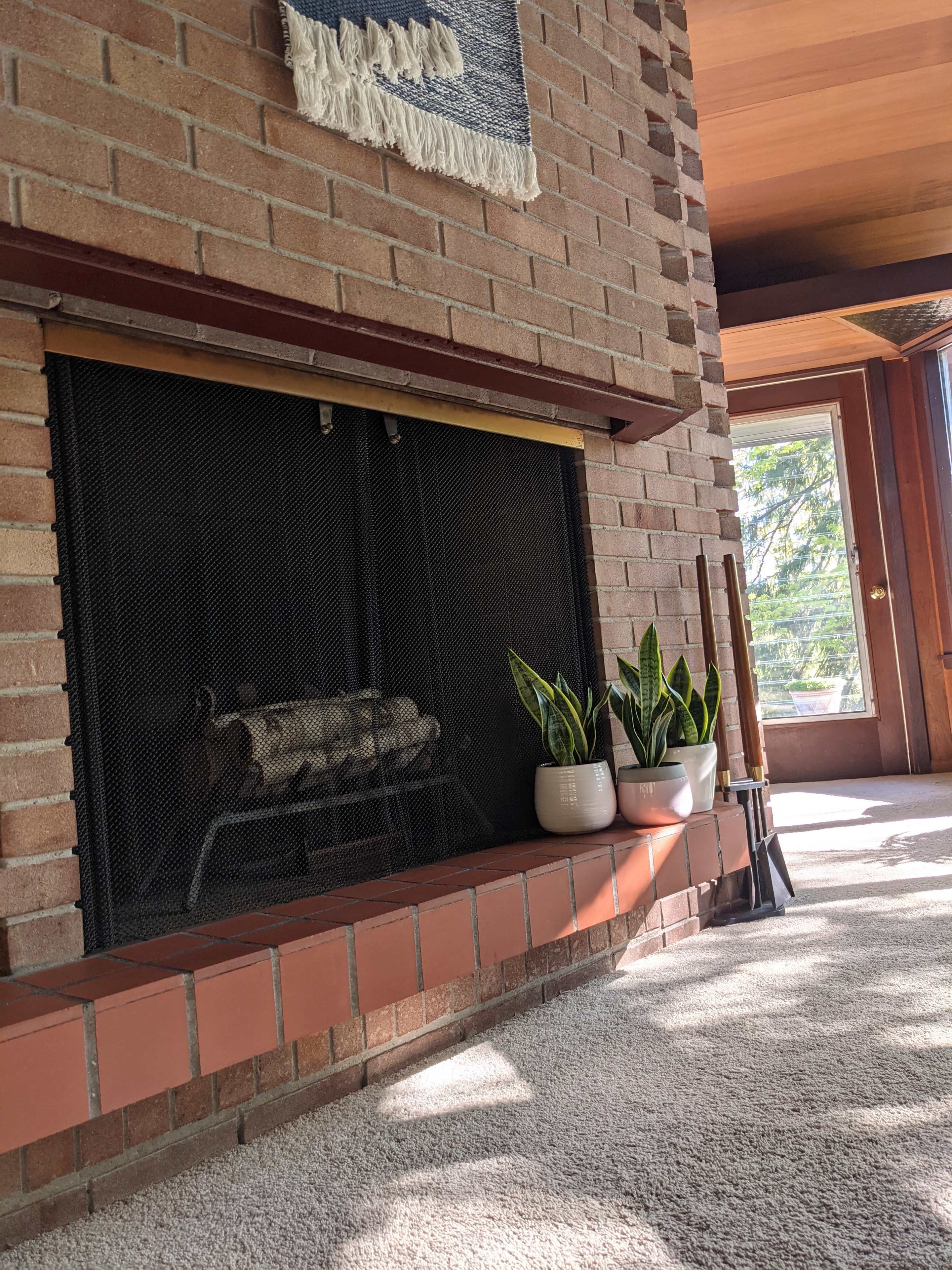 A brick fireplace with a mesh screen, accompanied by houseplants in white pots and a view of natural light coming from a window.