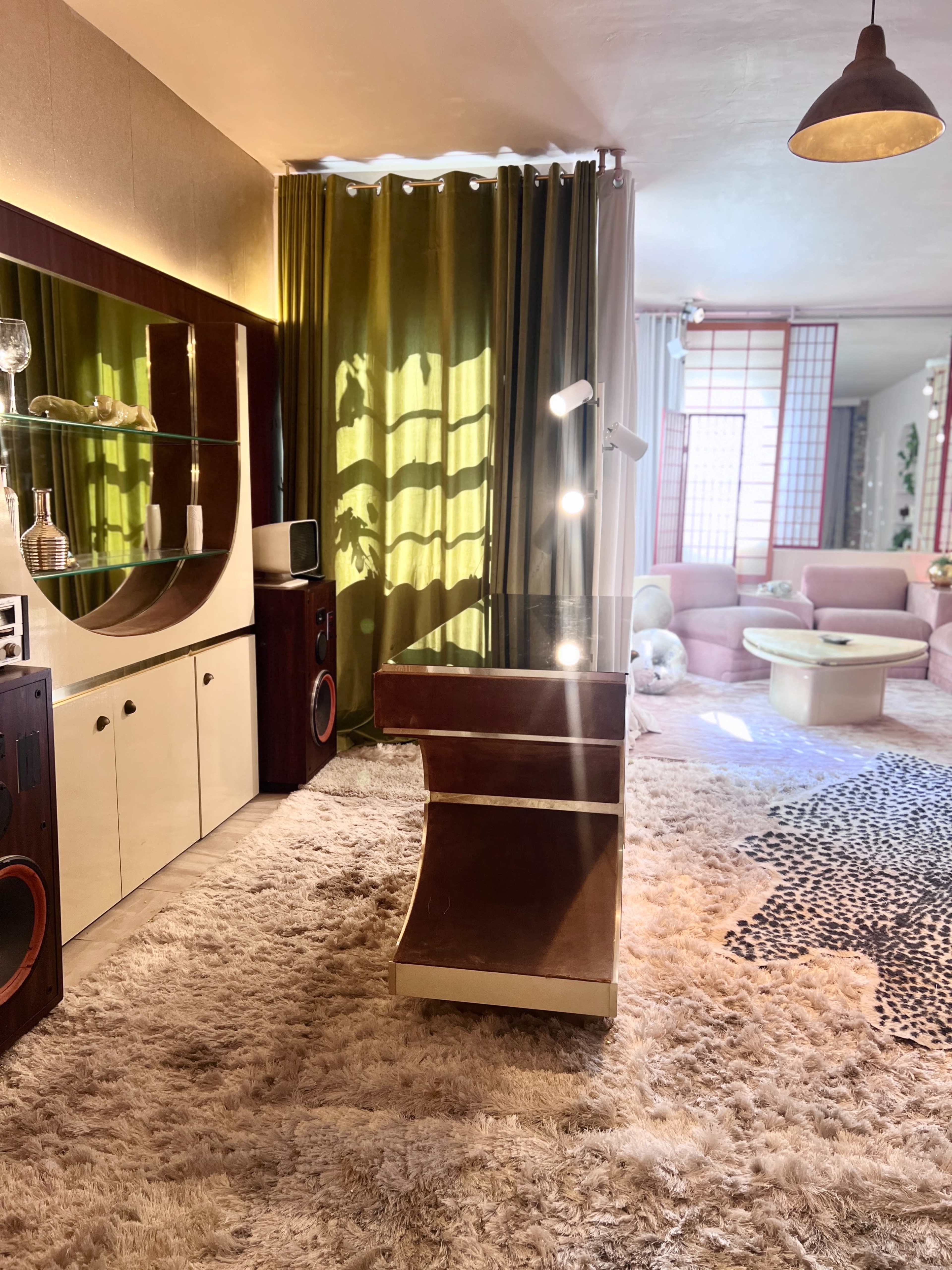 A stylish interior featuring a curved desk, large mirror, and a cozy seating area with pink furniture and green curtains.