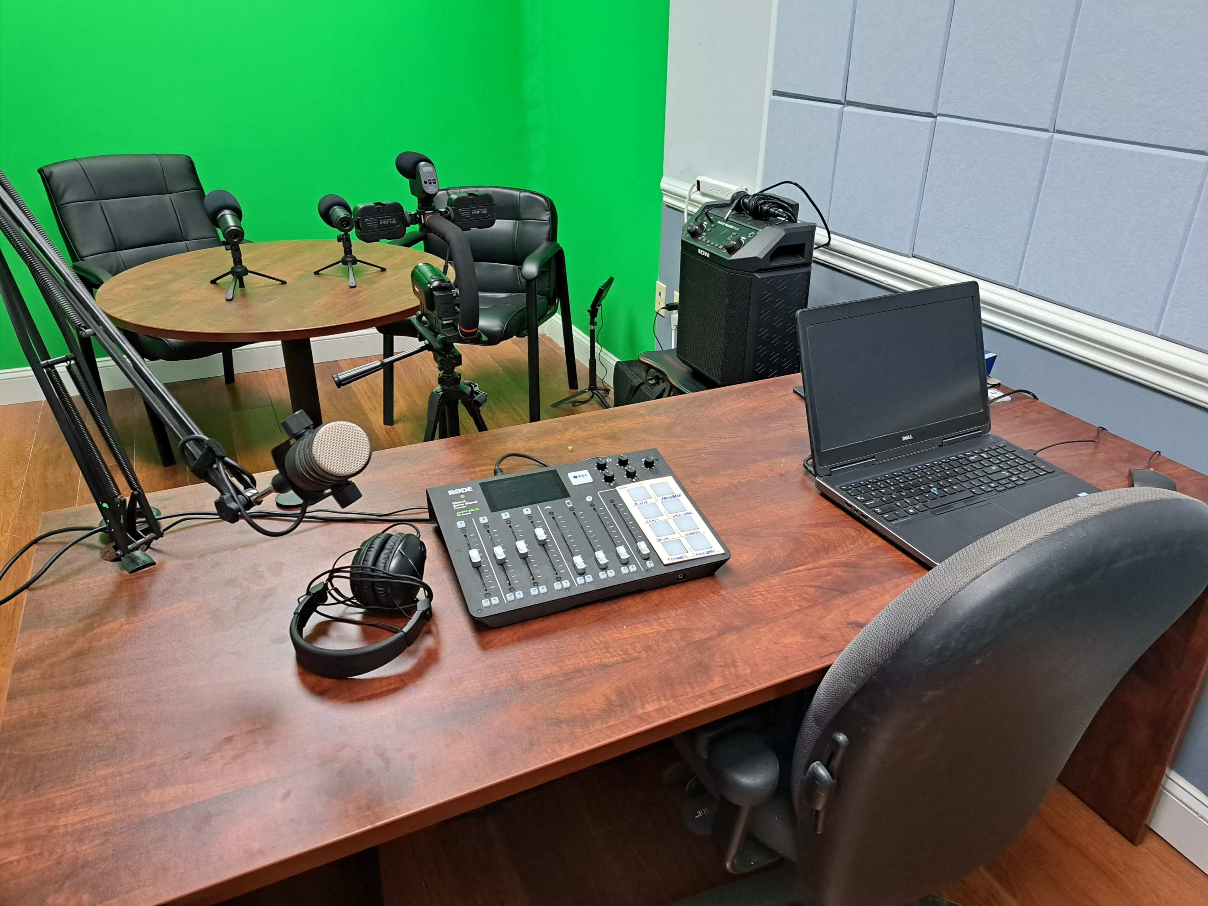 A recording studio with a green screen backdrop, microphones on stands, a mixing board, headphones, and a laptop on a wooden desk.