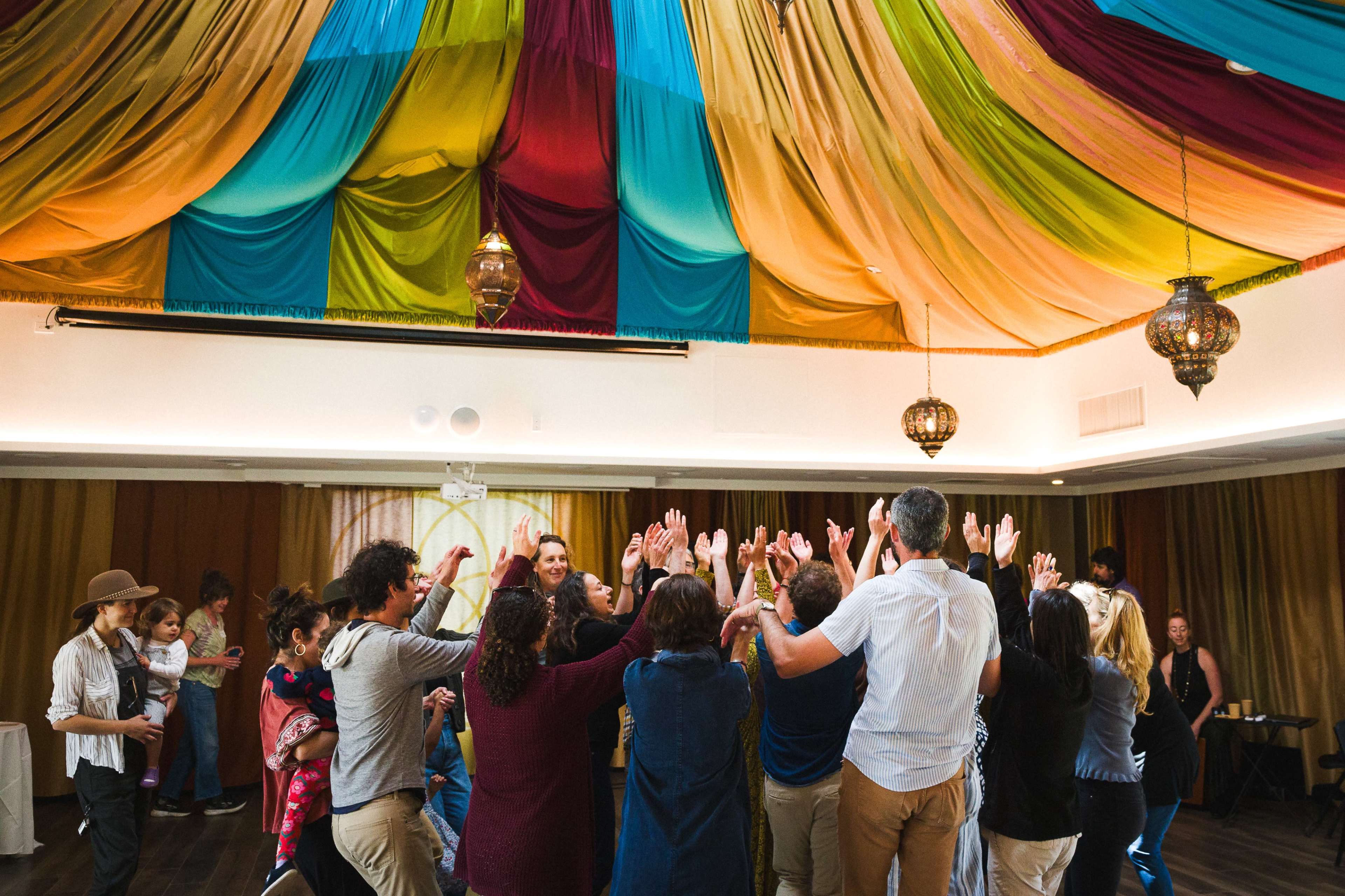 A group of people gather in a colorful room with draped fabric overhead, engaging in a lively activity while raising their hands.