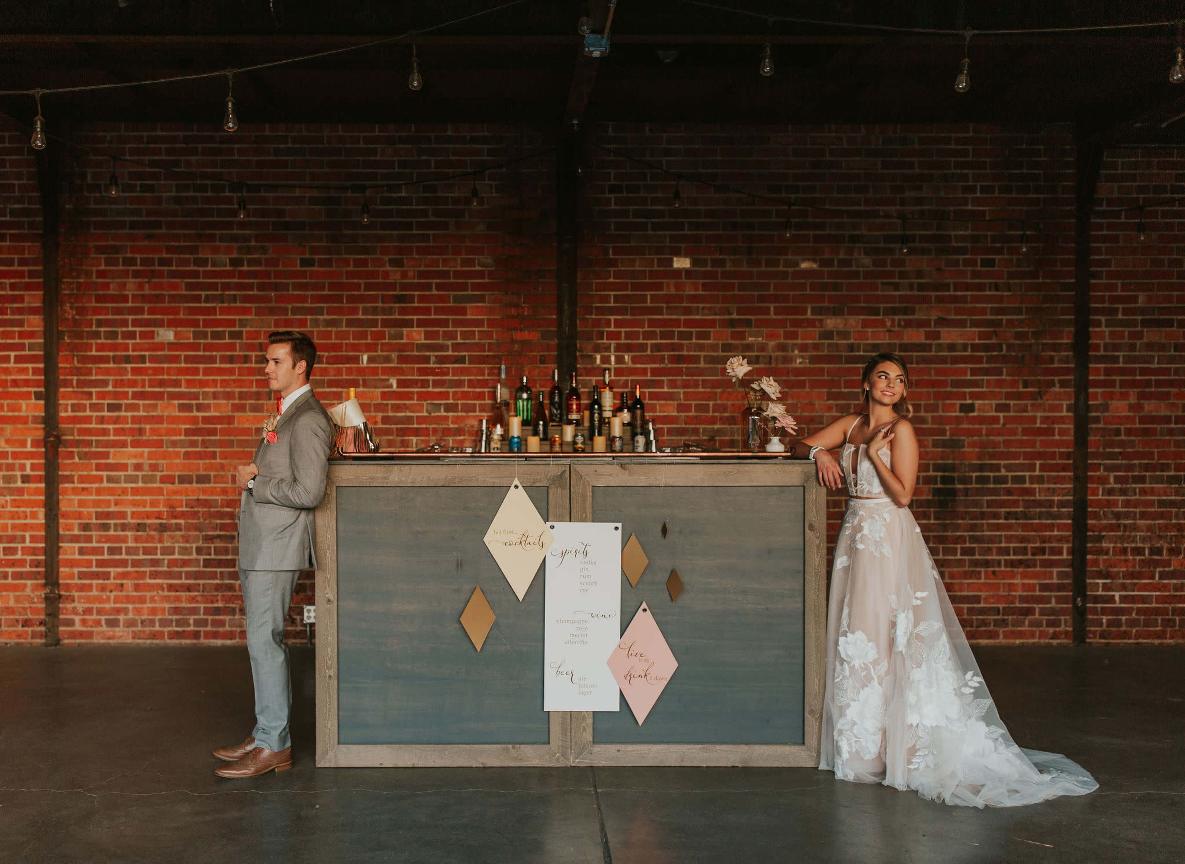 A couple stands on opposite sides of a vintage bar against a brick wall, with wedding decor displayed in front of them.