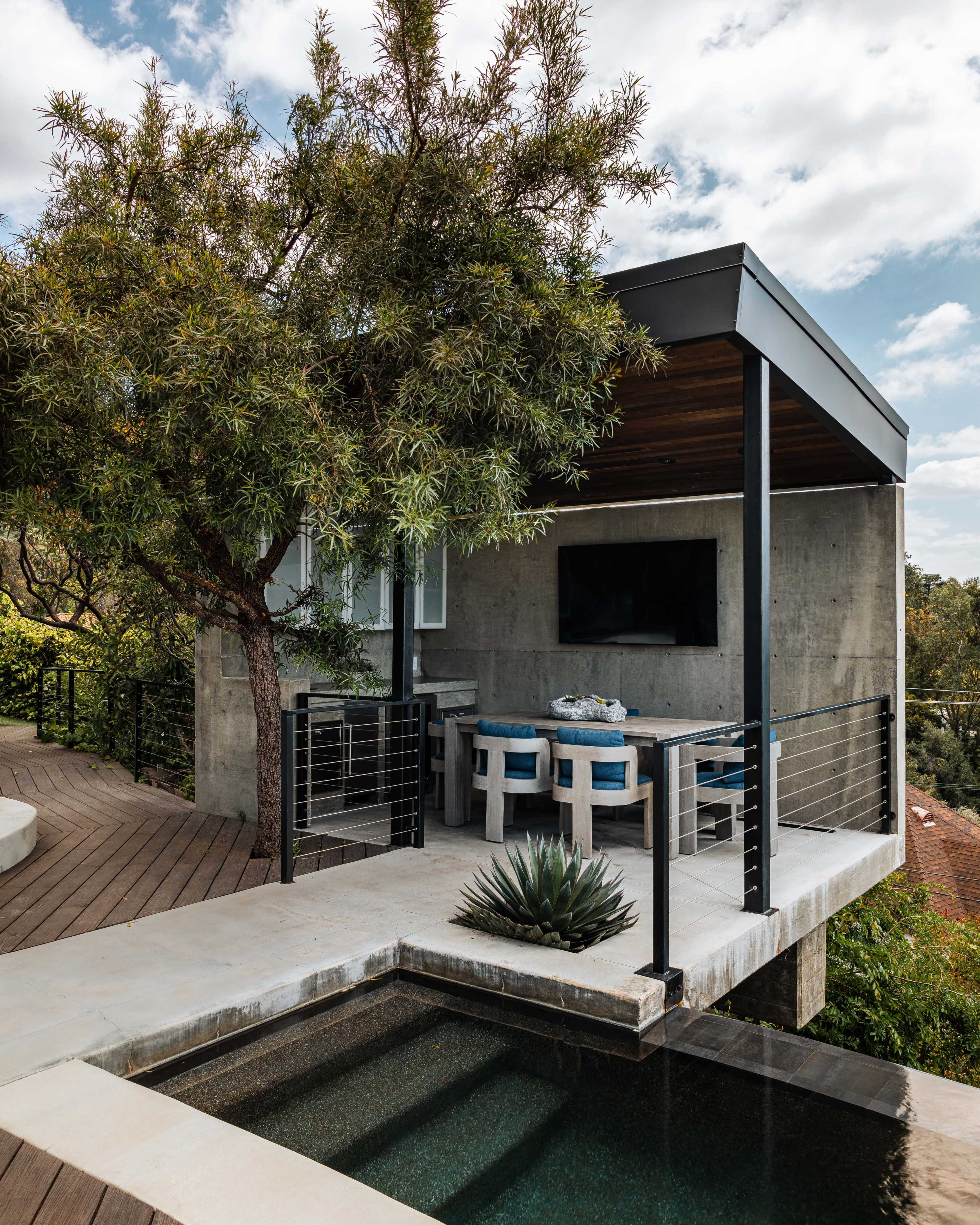A modern outdoor patio features a concrete structure with seating, a television, and a small swimming pool surrounded by greenery.