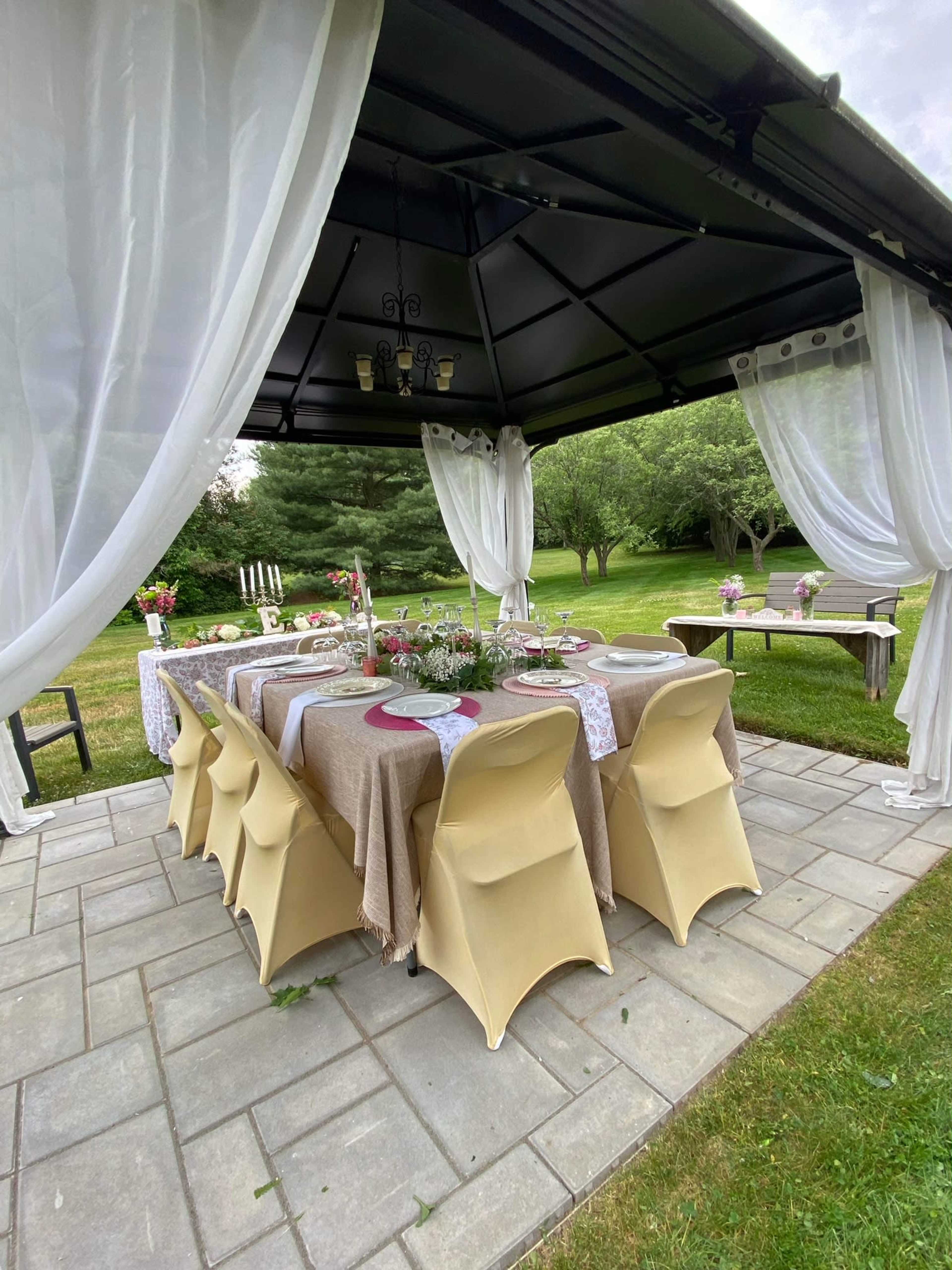 A covered outdoor dining area features a long table set with tableware and decorations, surrounded by grass and trees.