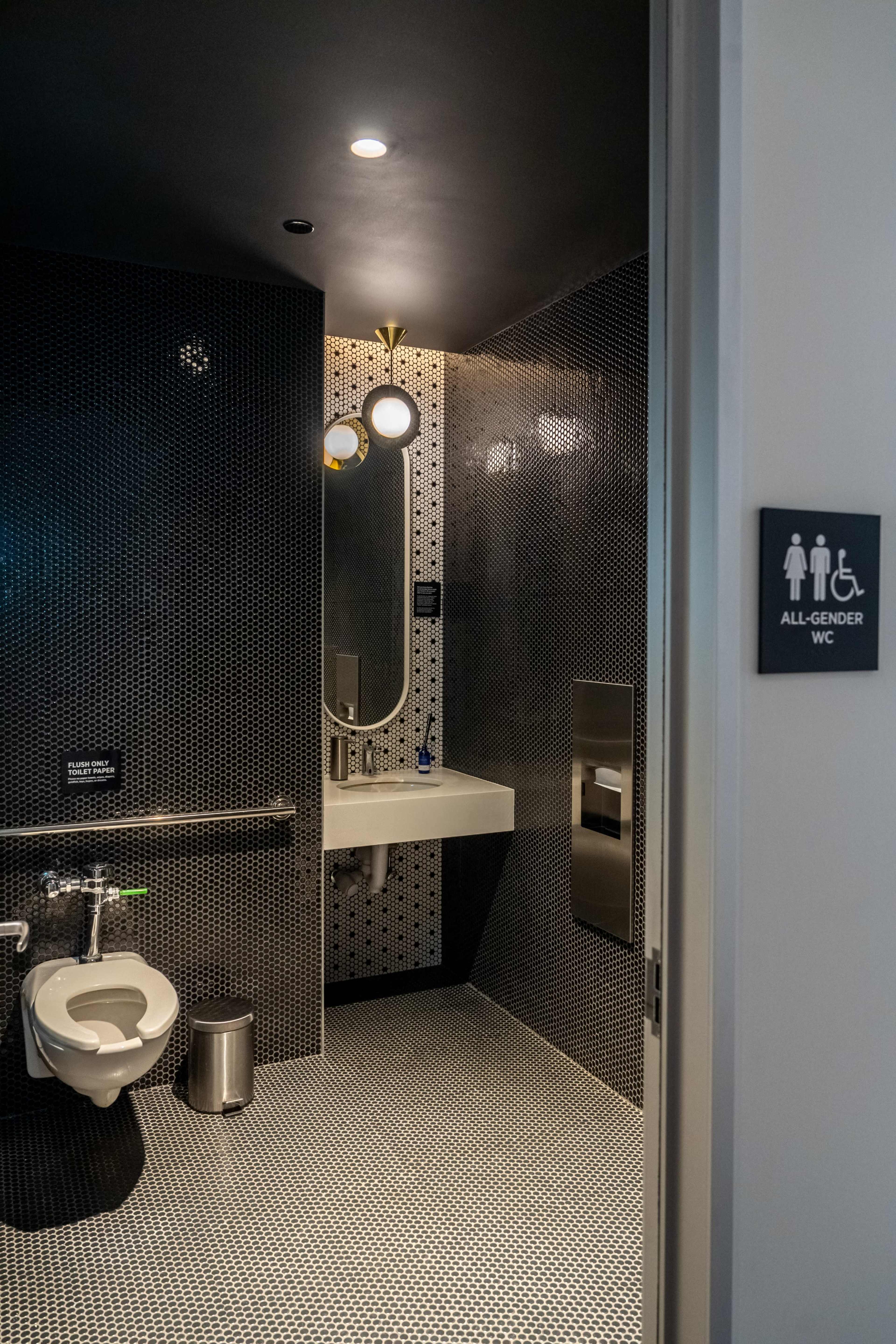 The image shows the entrance to a modern all-gender restroom featuring black tiled walls and a minimalist design, including a single toilet and a sink.