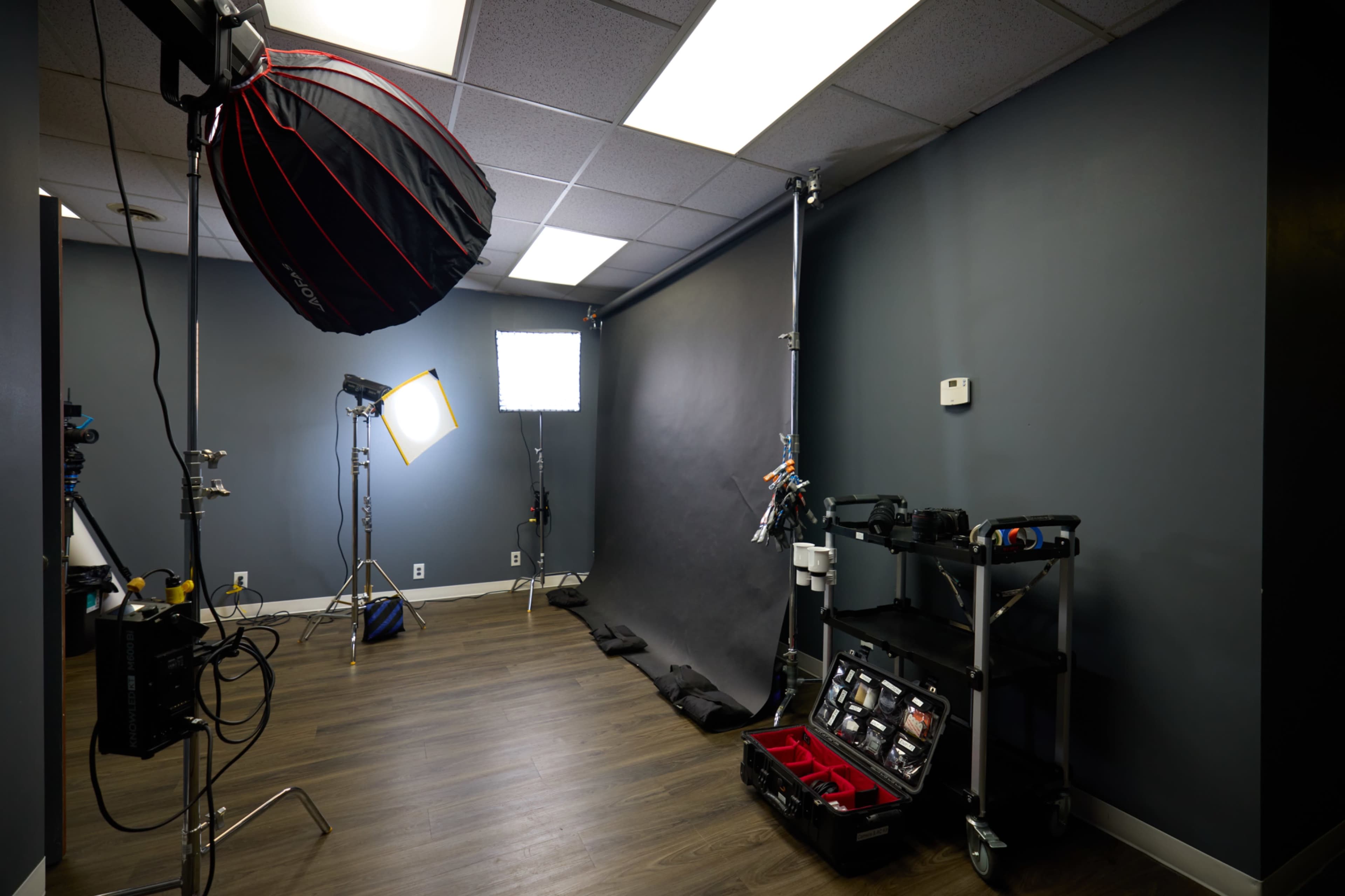 The image shows a photography studio with a black backdrop, lighting equipment, and a cart containing various tools and accessories.