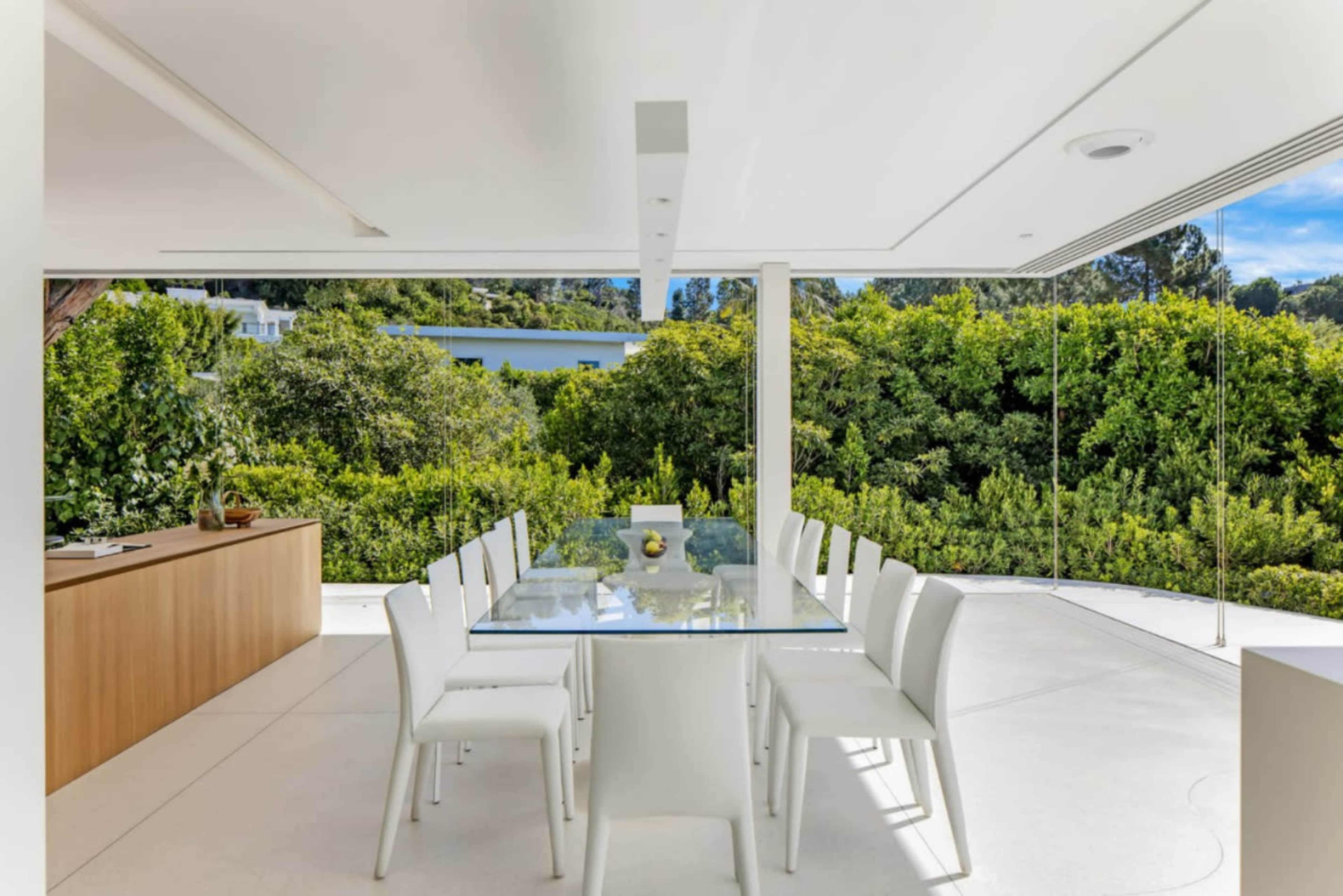 The image shows a modern dining area with a glass table surrounded by white chairs, adjacent to a wall of greenery.
