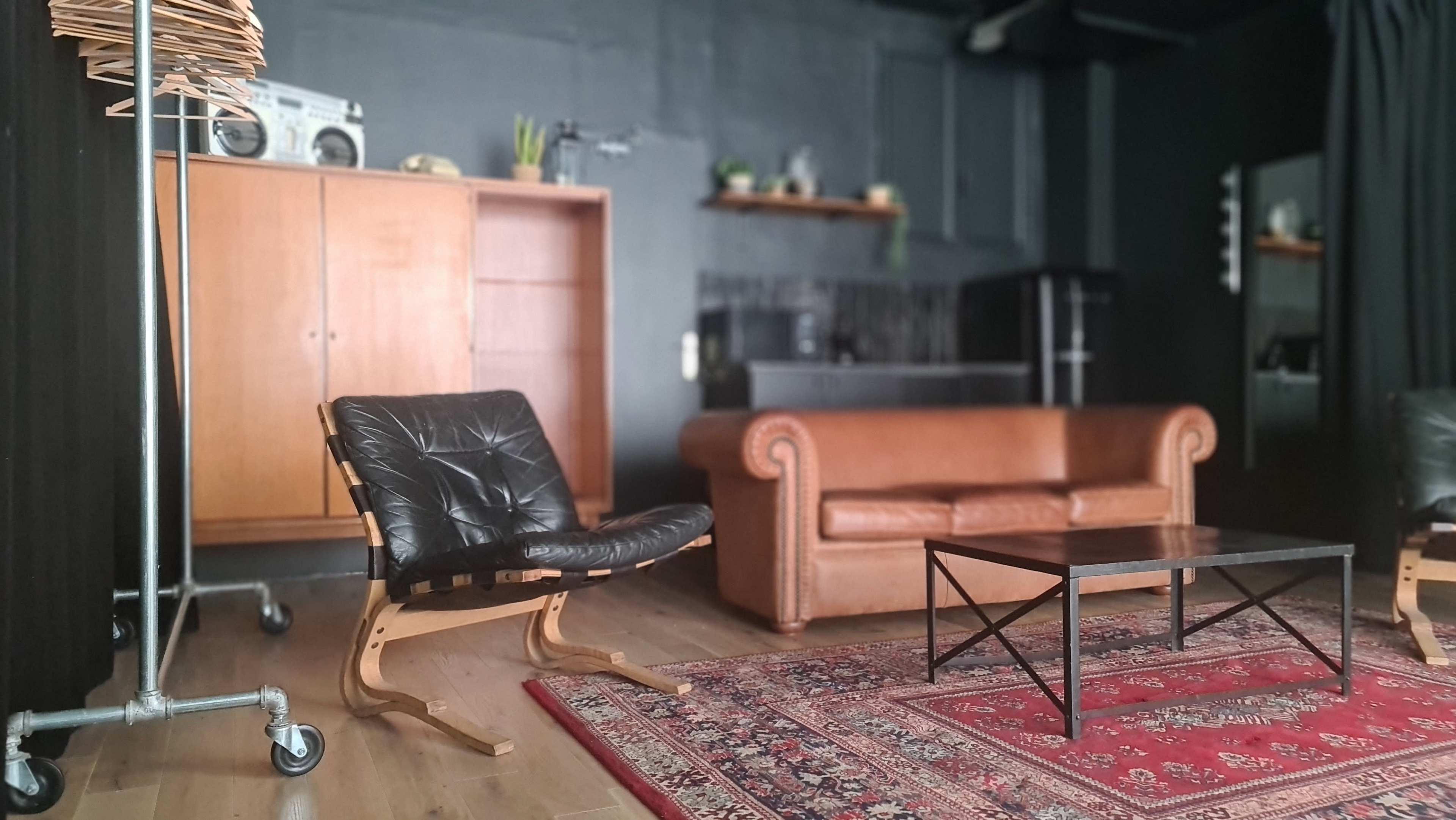 The image shows a stylish interior featuring a brown leather couch, a black chair, a coffee table, a patterned rug, and wooden furniture against a dark wall.