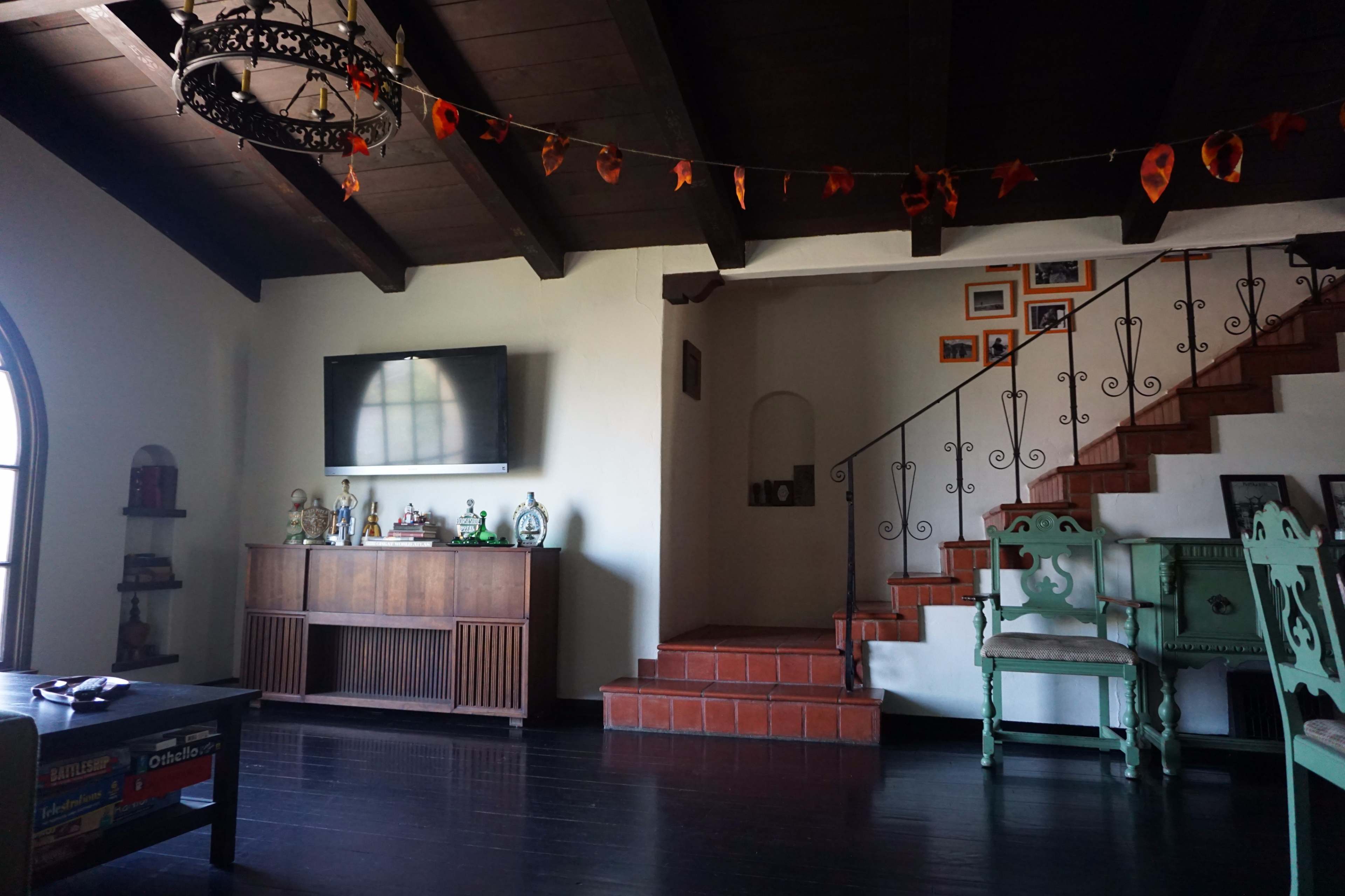 The image shows a living room with a wooden ceiling, a television mounted on the wall, and a staircase leading to an upper level, accompanied by decorative items and a green chair.