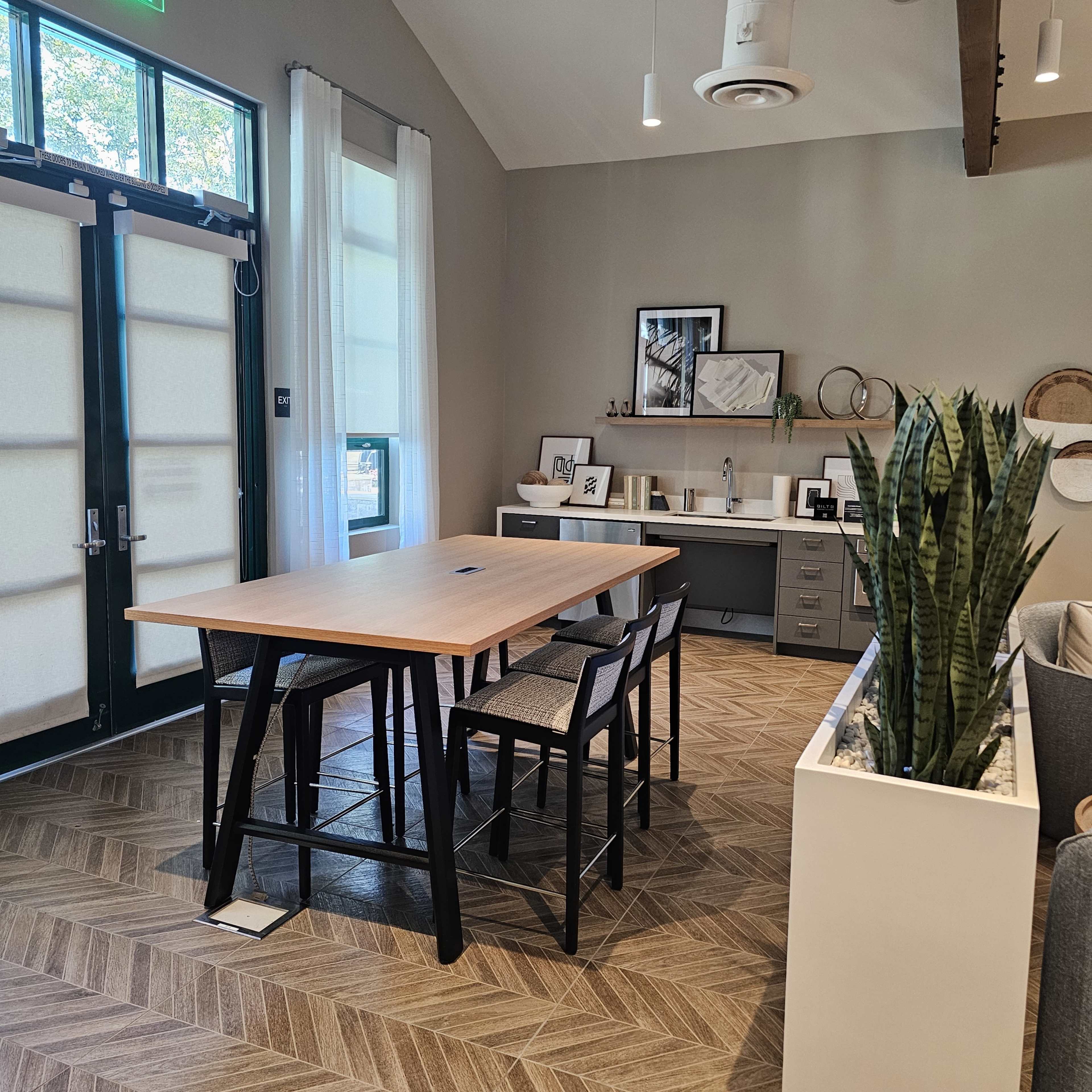 A modern, light-filled room with a large wooden table and chairs, a kitchenette area, and a decorative plant in a white planter.