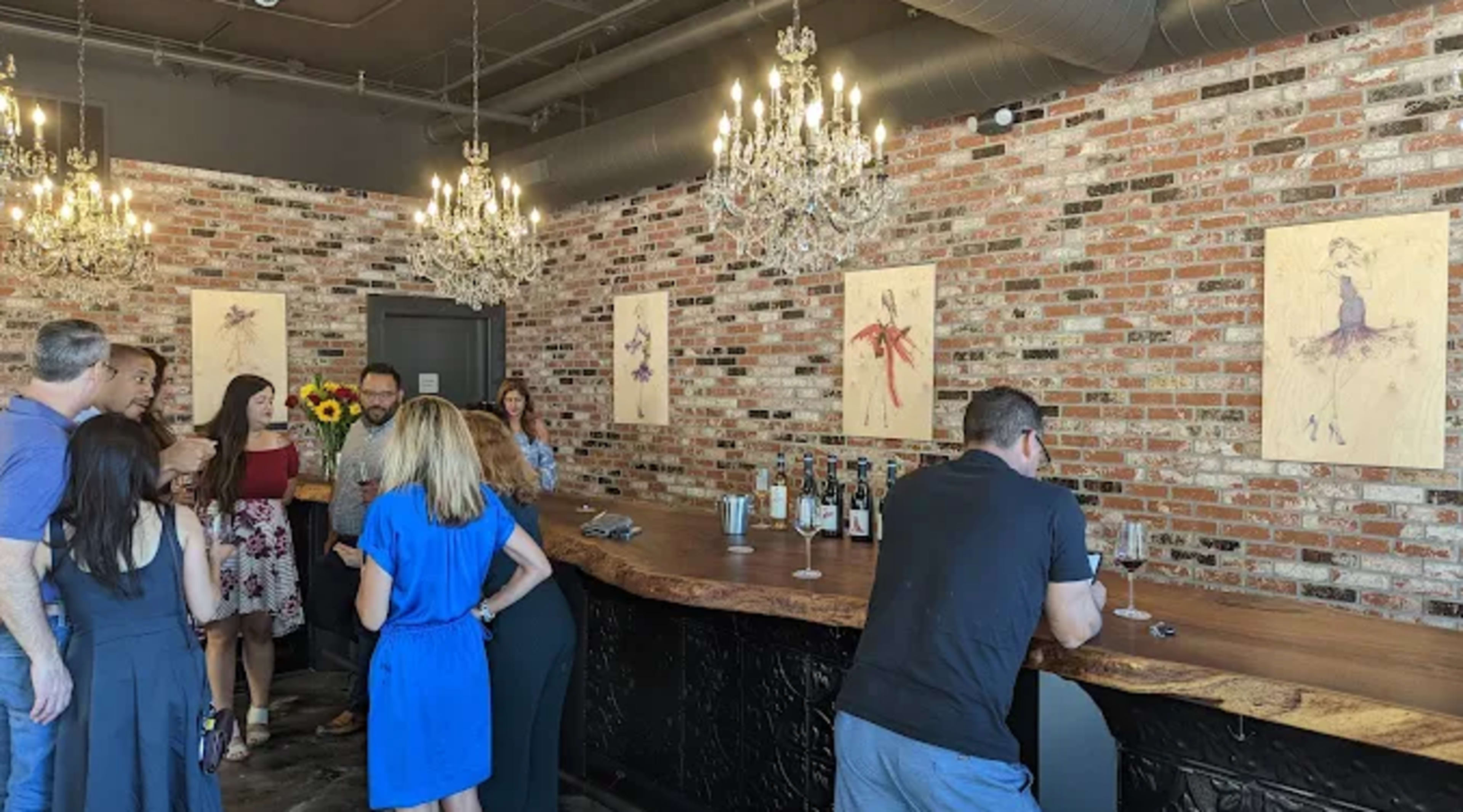 A group of people mingles in a stylish room with a brick wall adorned with artwork, chandeliers overhead, and a long wooden bar featuring bottles of wine.