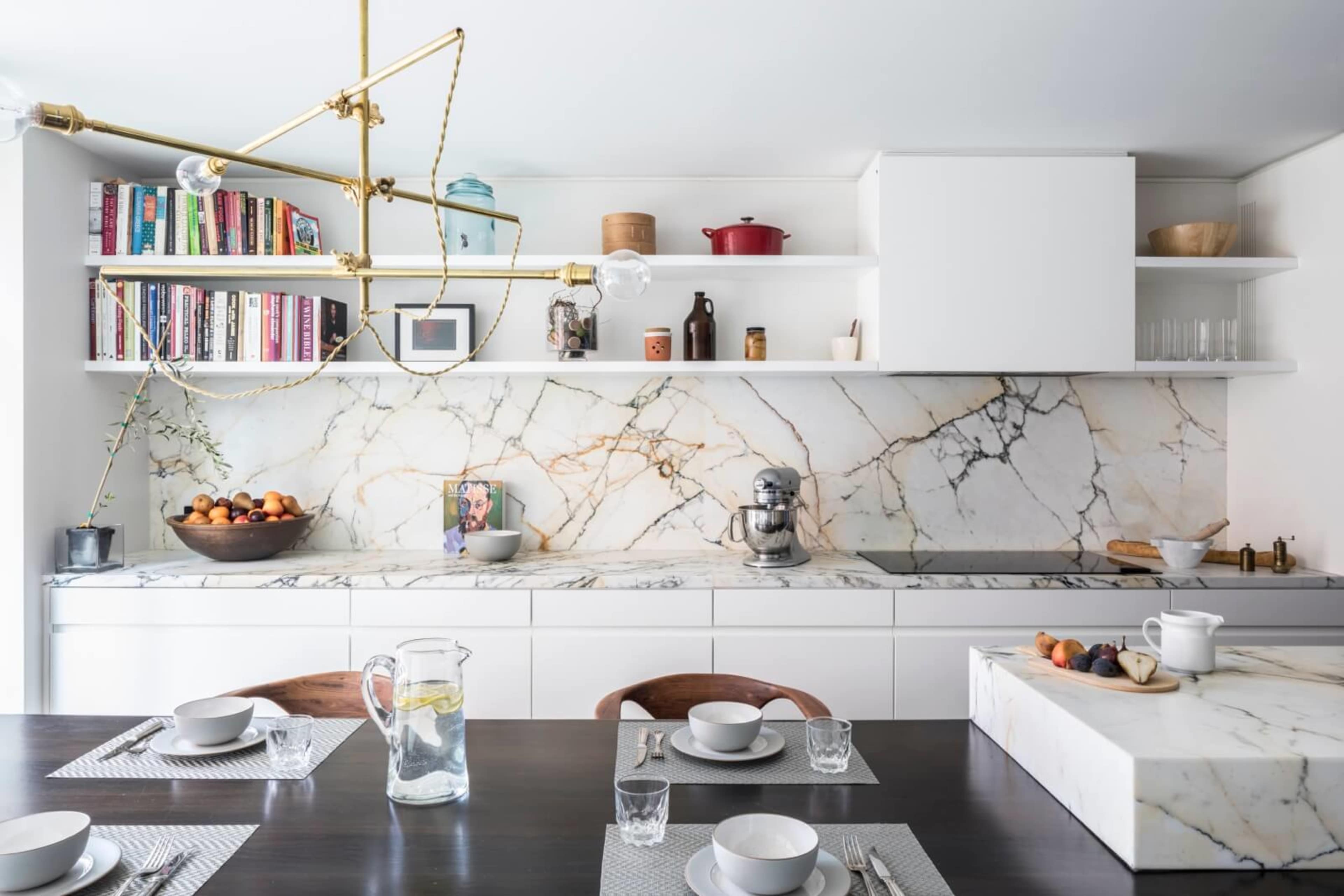 A modern kitchen features a marble backsplash, white cabinetry, open shelving with books, and a dining table set with dishes and glassware.