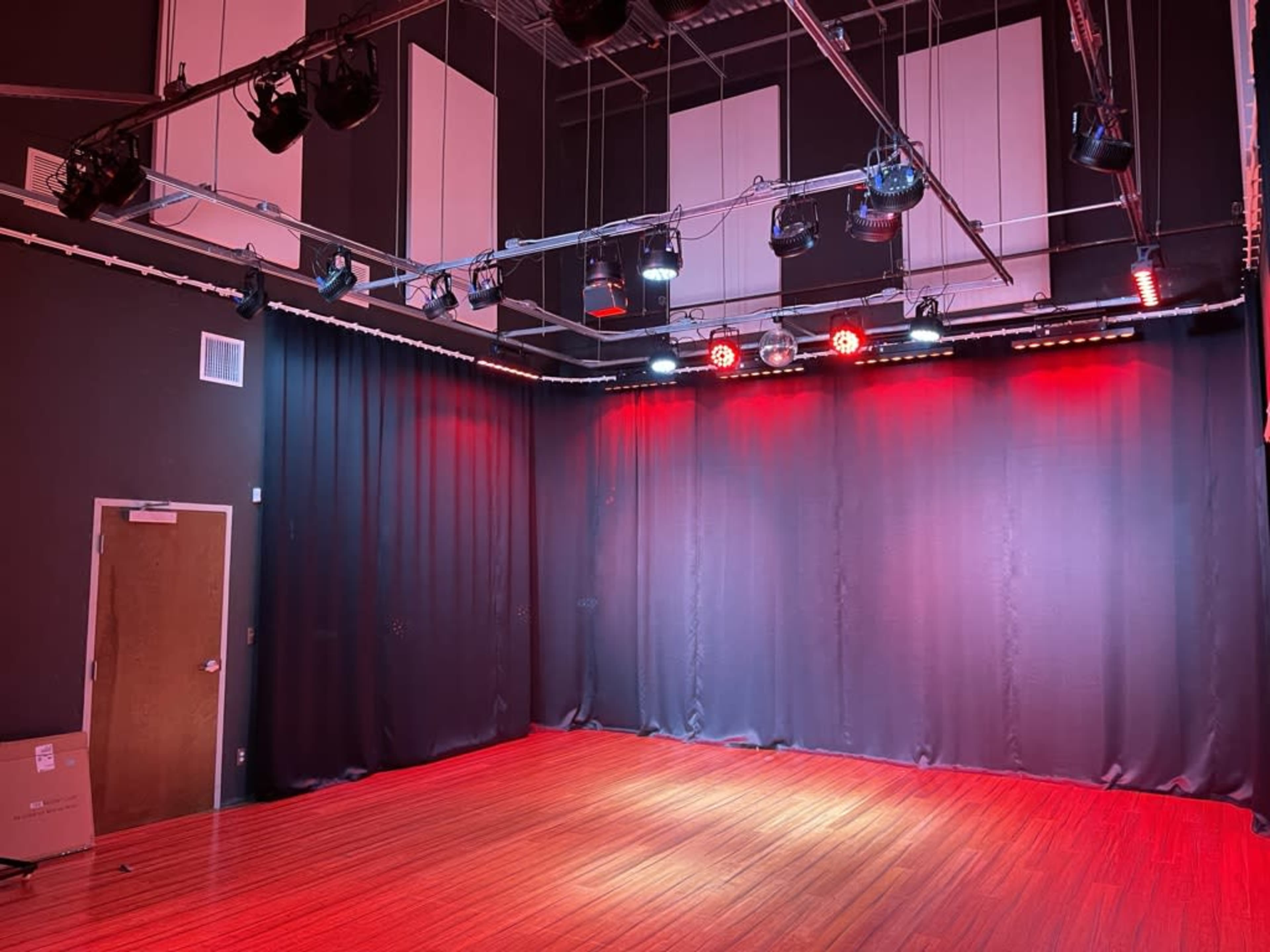 A dimly lit performance space with wooden flooring, black curtains on the sides, and overhead stage lights in red and white.