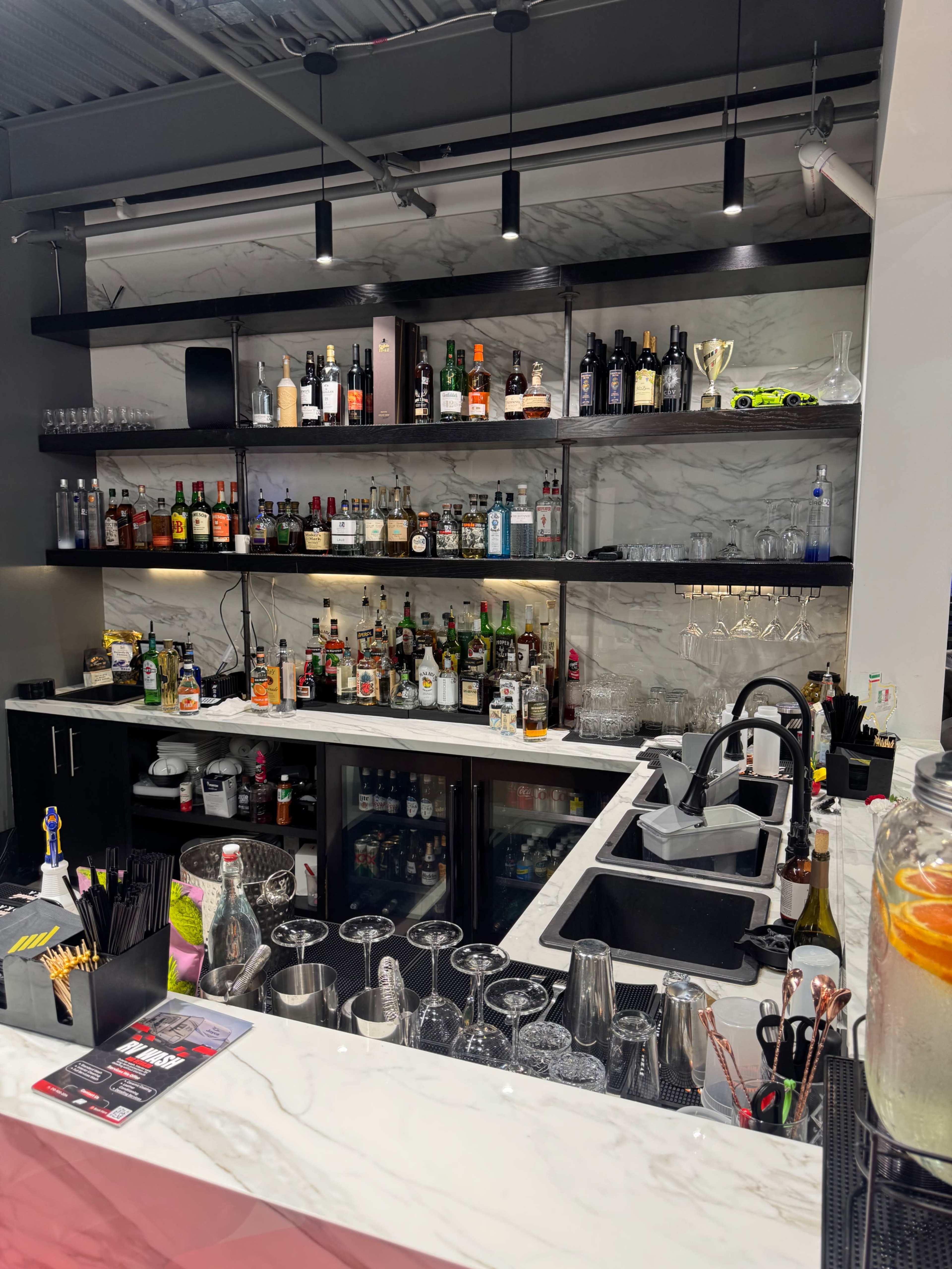 The image shows a well-stocked bar area with shelves holding various bottles of liquor and glassware, alongside a clean countertop with cocktail-making tools.