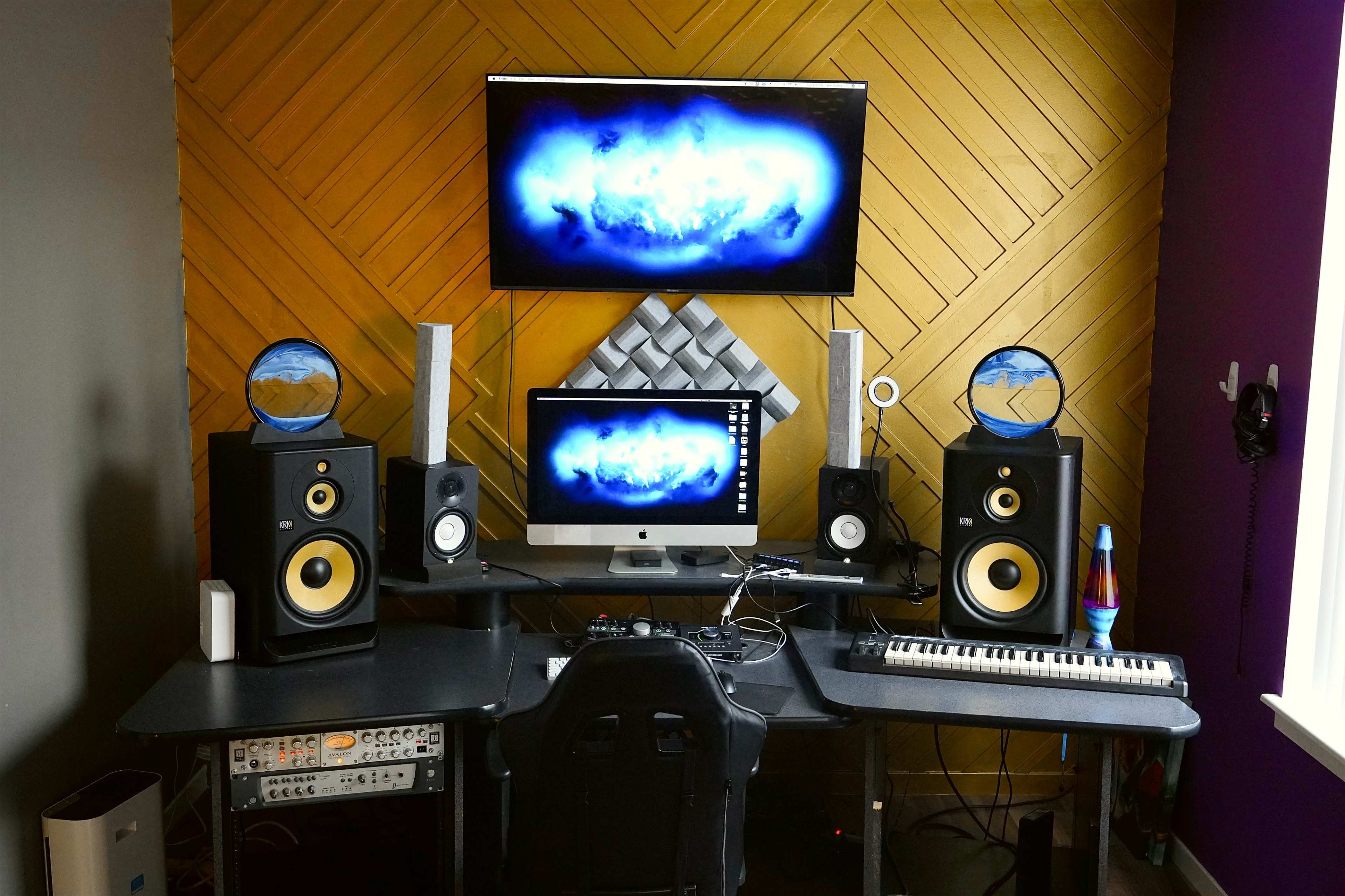 The image shows a music production studio featuring a desk with a computer, audio monitors, a MIDI keyboard, and a television mounted on the wall above.