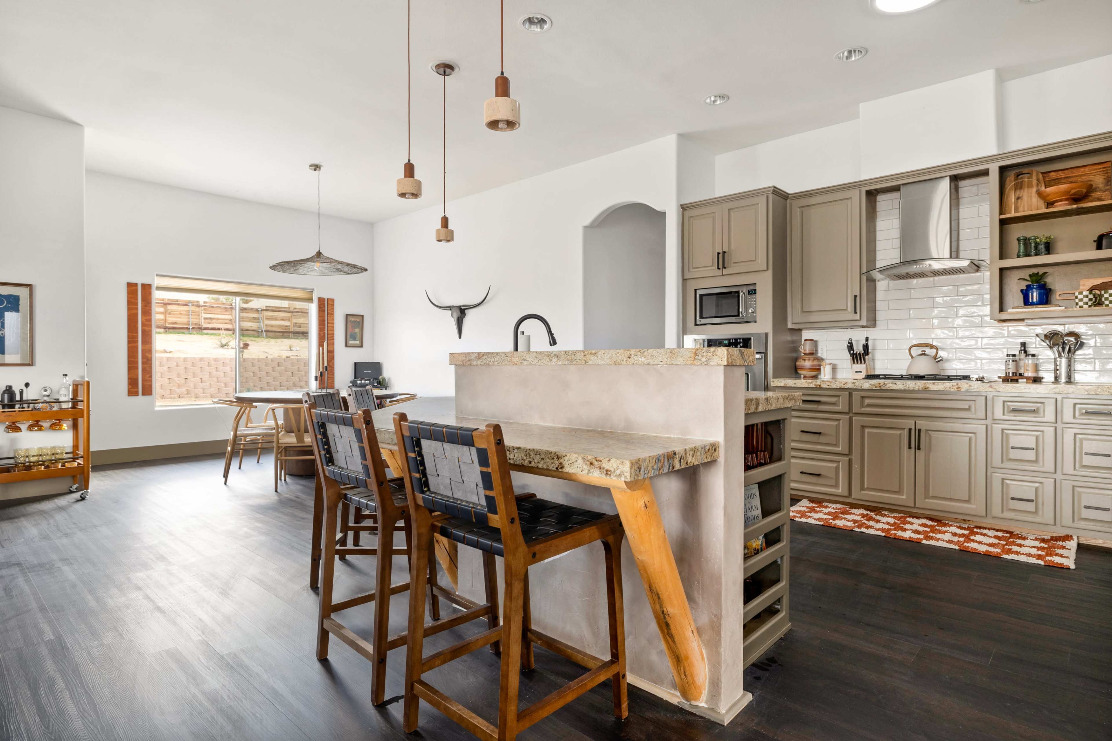 The image shows a modern kitchen with a breakfast bar, featuring wooden stools, stainless steel appliances, and a combination of light and dark cabinetry.