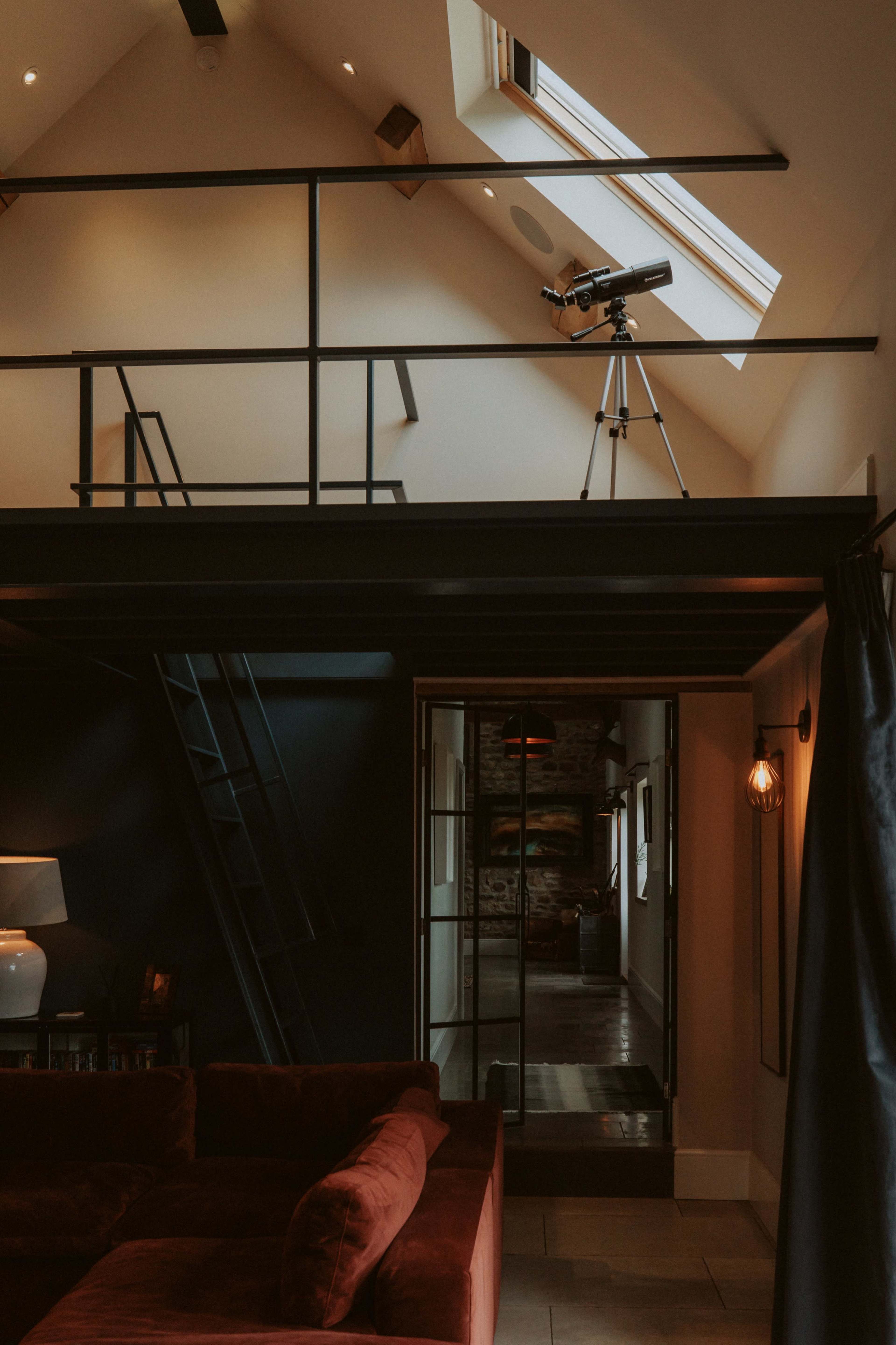 A telescope is positioned near a skylight on a lofted area overlooking a cozy living space with a sectional sofa and modern decor.
