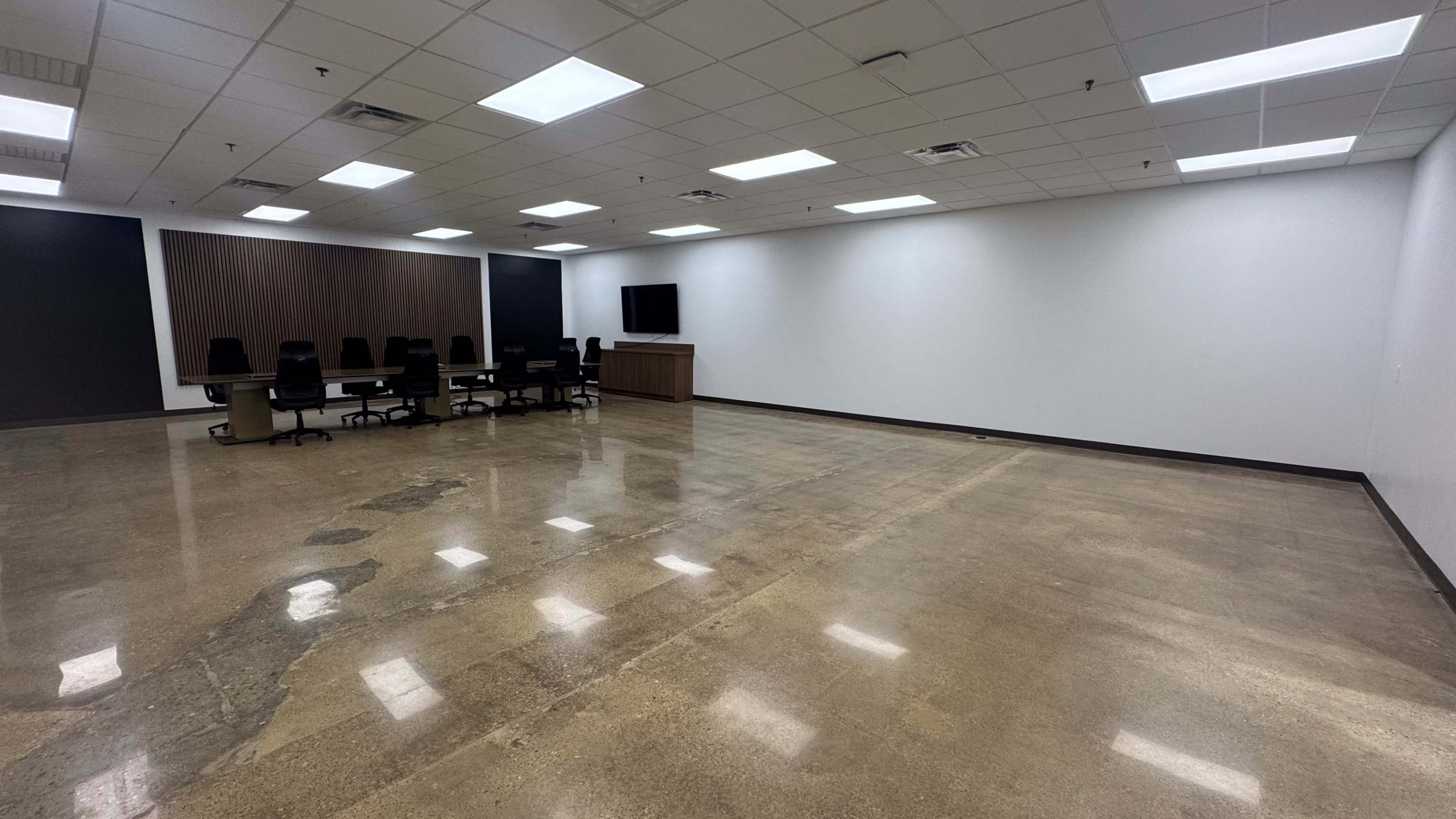 The image shows a spacious, empty room with polished concrete floors, a conference table, chairs, and a wall-mounted television.