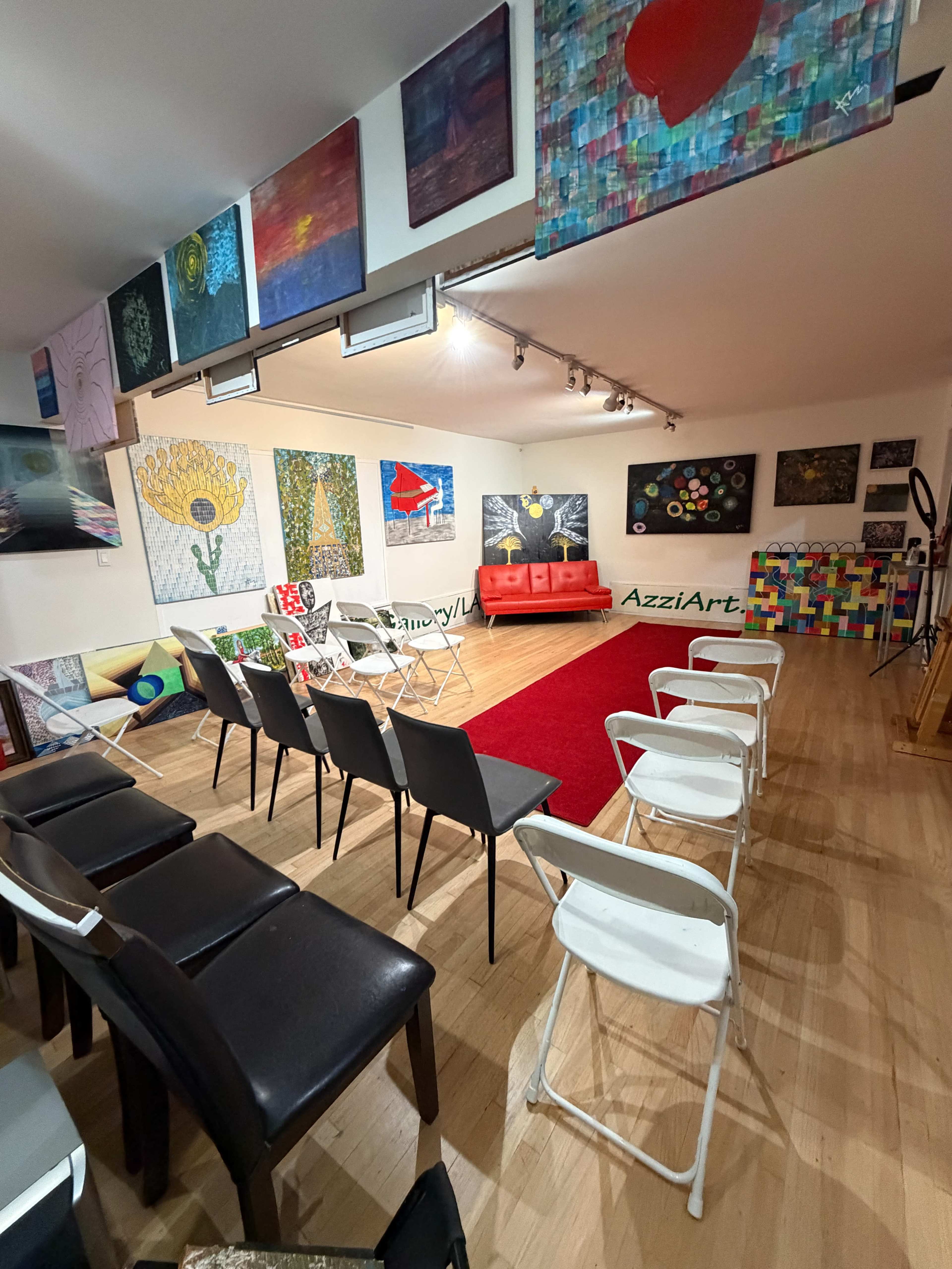 The image shows an art gallery or studio with rows of white and black chairs arranged facing a red carpet and several colorful artworks displayed on the walls.