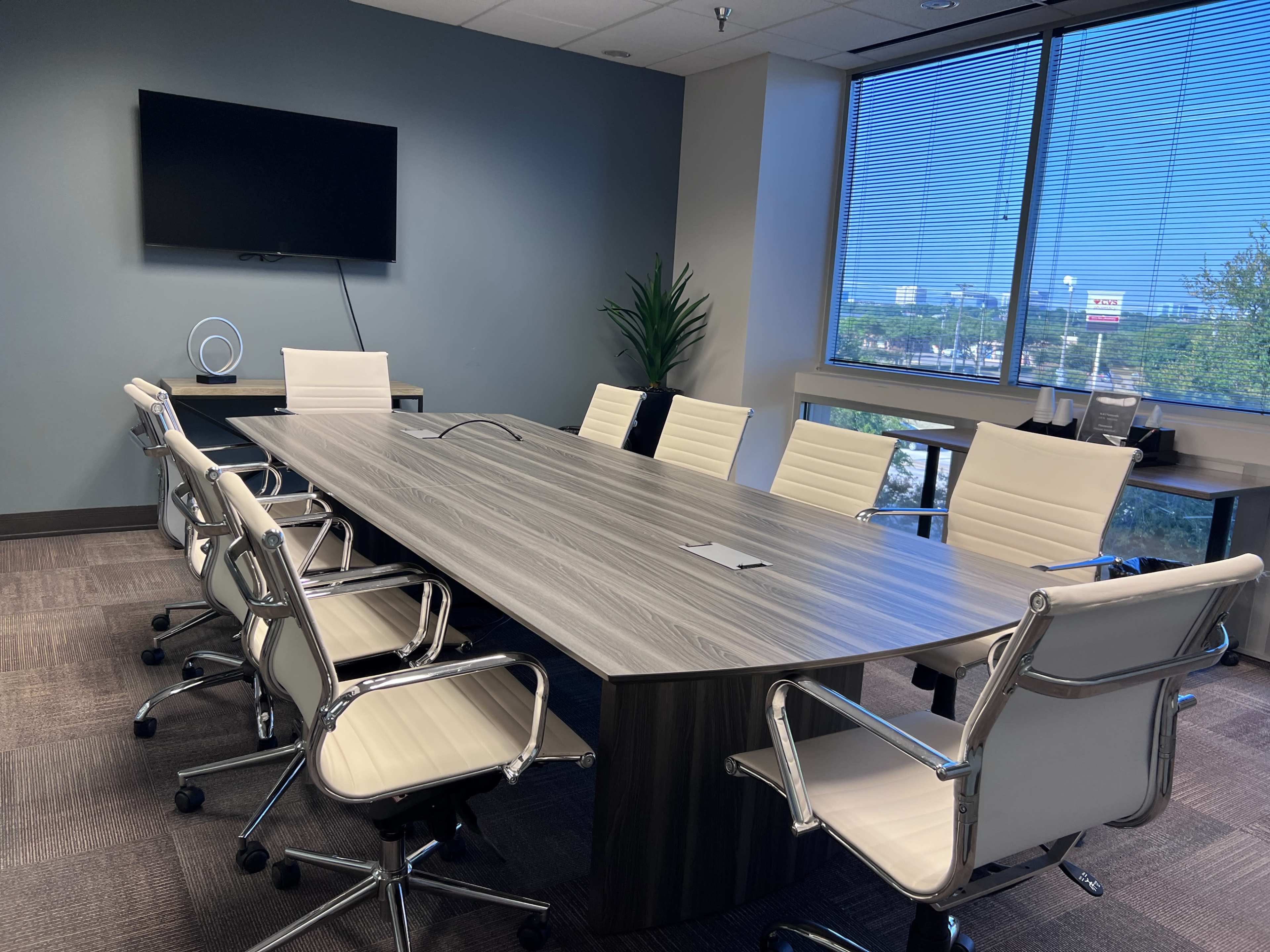 A modern conference room features a long wooden table surrounded by eight white leather chairs, with a television mounted on the wall and large windows overlooking a cityscape.