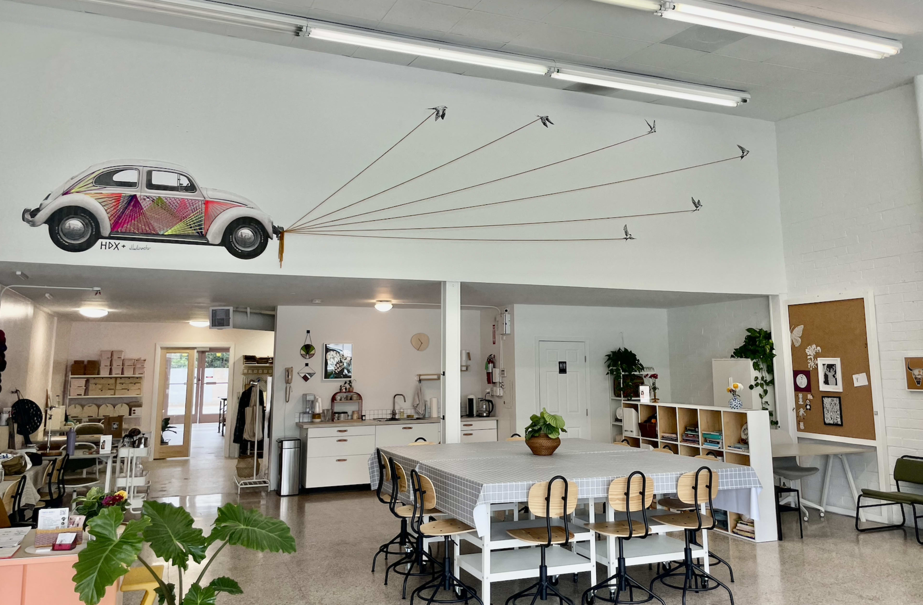 The interior of a modern café features a long communal table, a mural of a colorful car on the wall, and decorative plants scattered around.