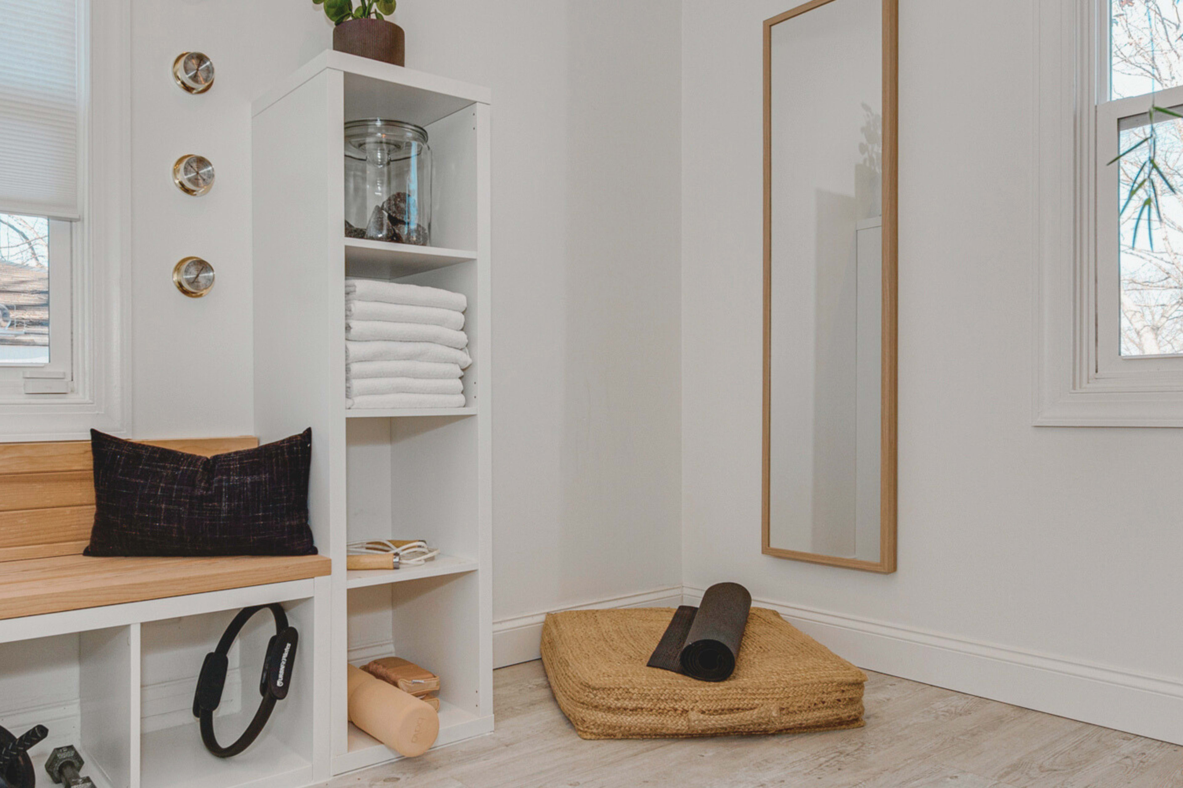 A bright room featuring a wooden bench, a white shelf with neatly stacked towels, a large mirror, and a rolled yoga mat on a woven mat.
