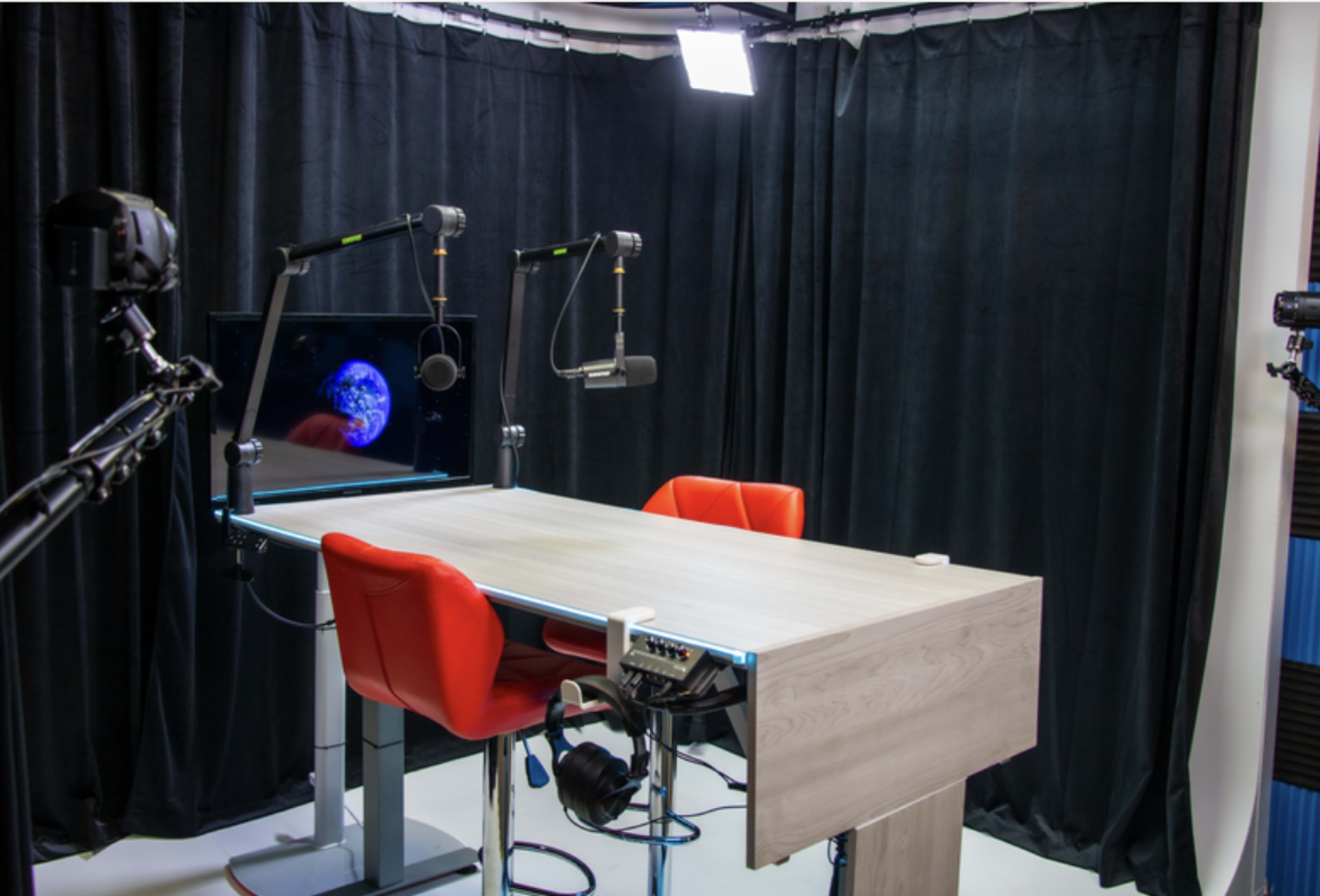 A modern podcasting studio features a wooden table, two red chairs, and microphones, with a screen displaying an image of Earth in the background.