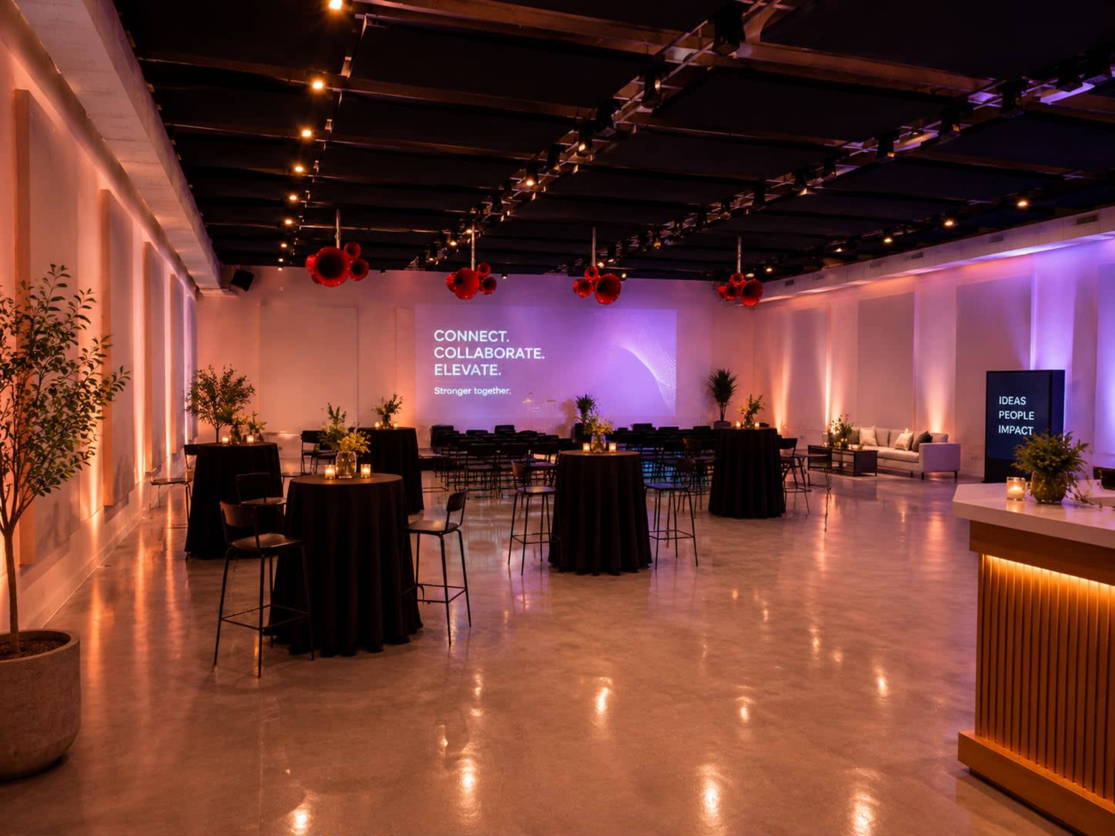 The image shows a brightly lit event space with black tables, plants, and a large projection screen displaying motivational phrases.