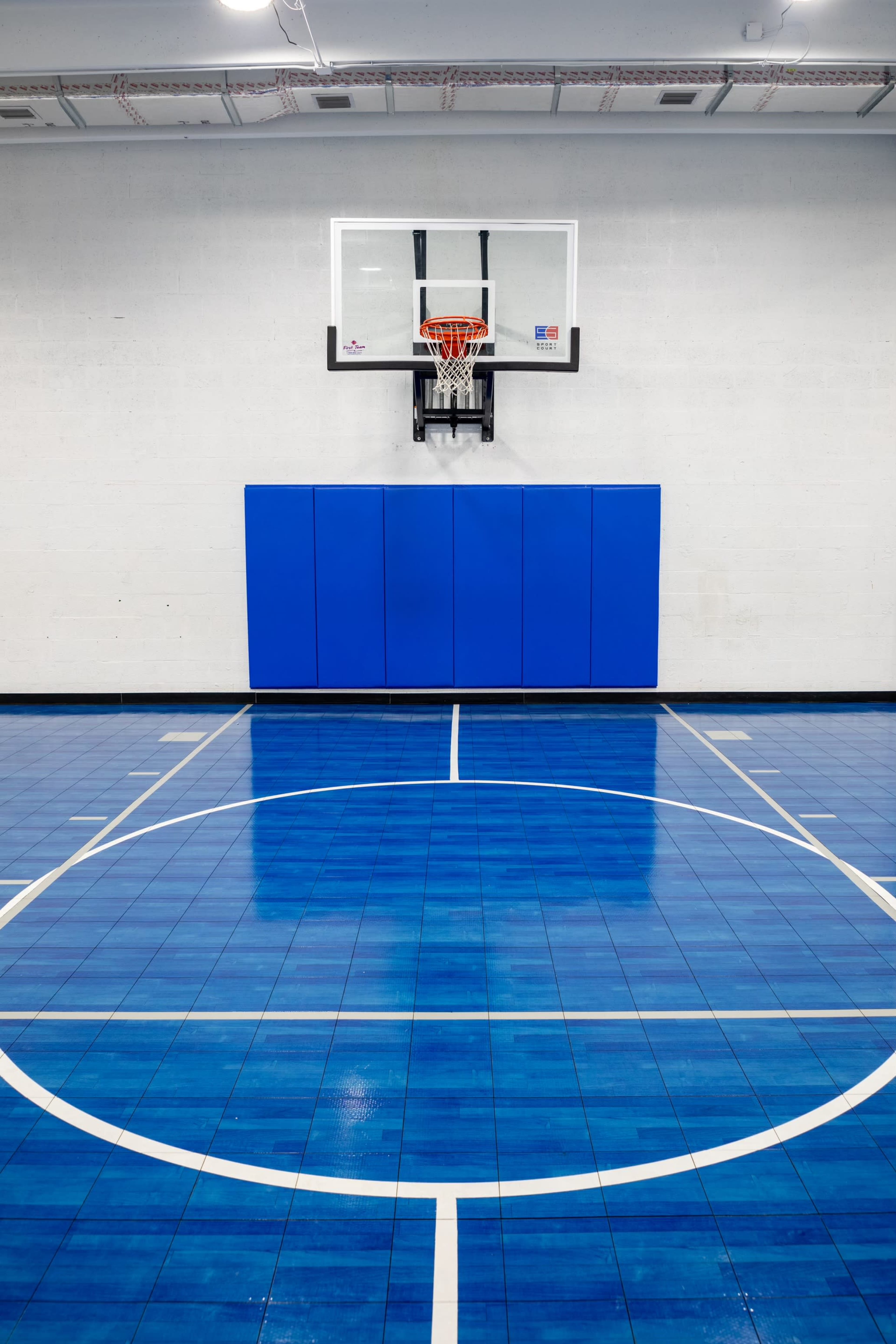 A basketball court with a blue and white patterned floor, a hoop mounted on the wall, and padded wall sections behind the hoop.
