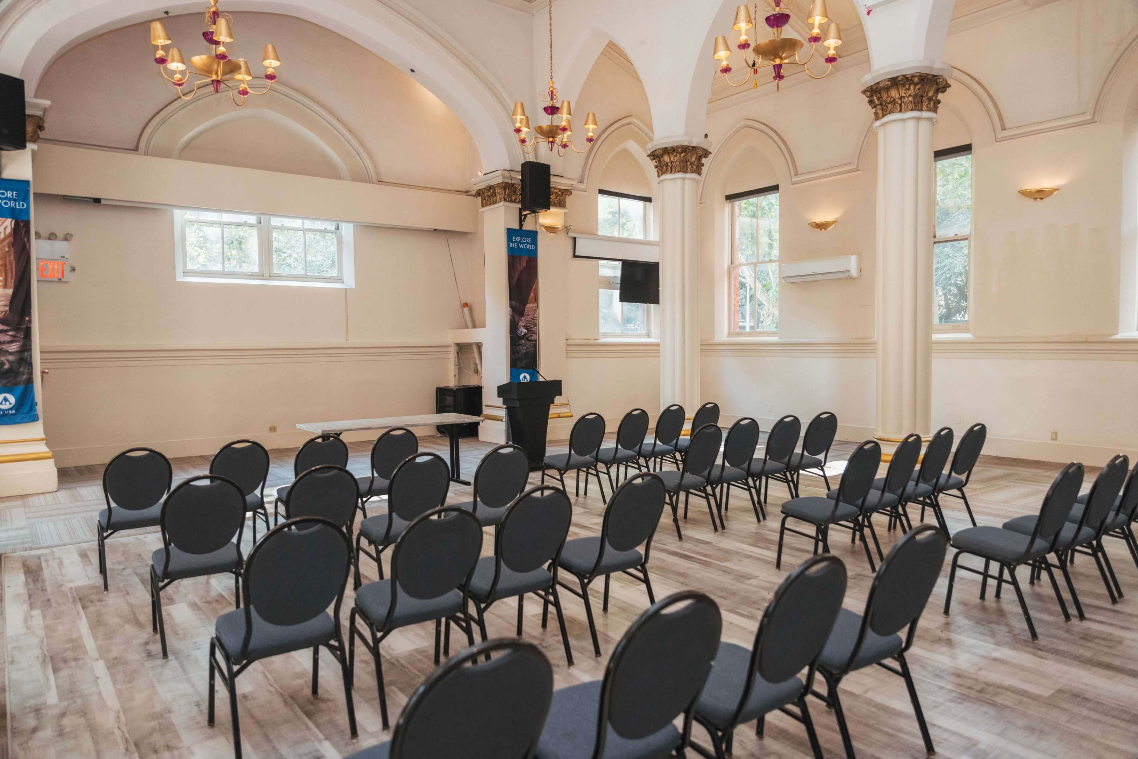 The image shows a spacious event room with rows of black chairs arranged for an assembly, adorned with ornate light fixtures and large arched windows.