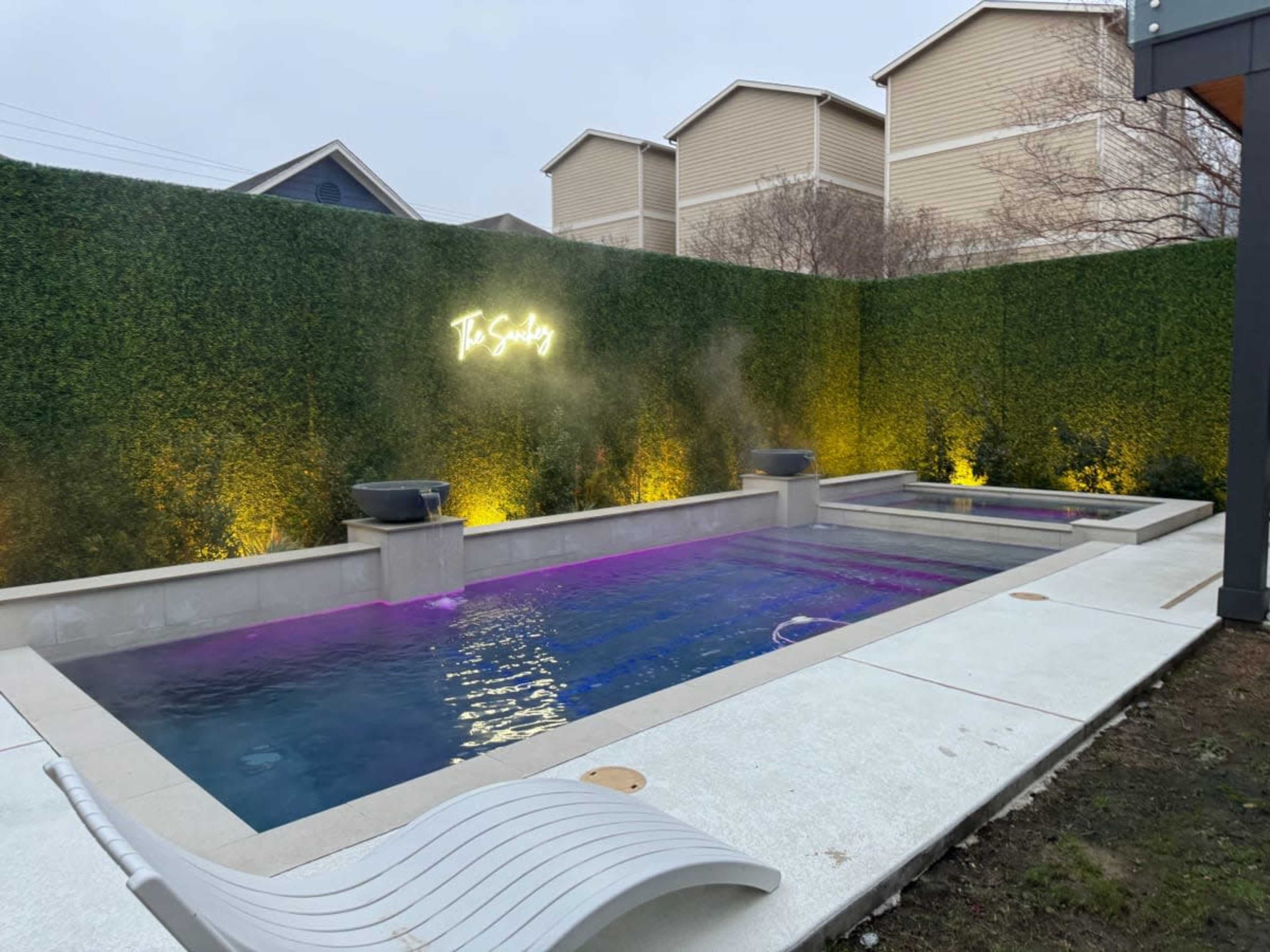 The image shows a modern outdoor pool area with two pools surrounded by a tall green hedge and illuminated by yellow lights.