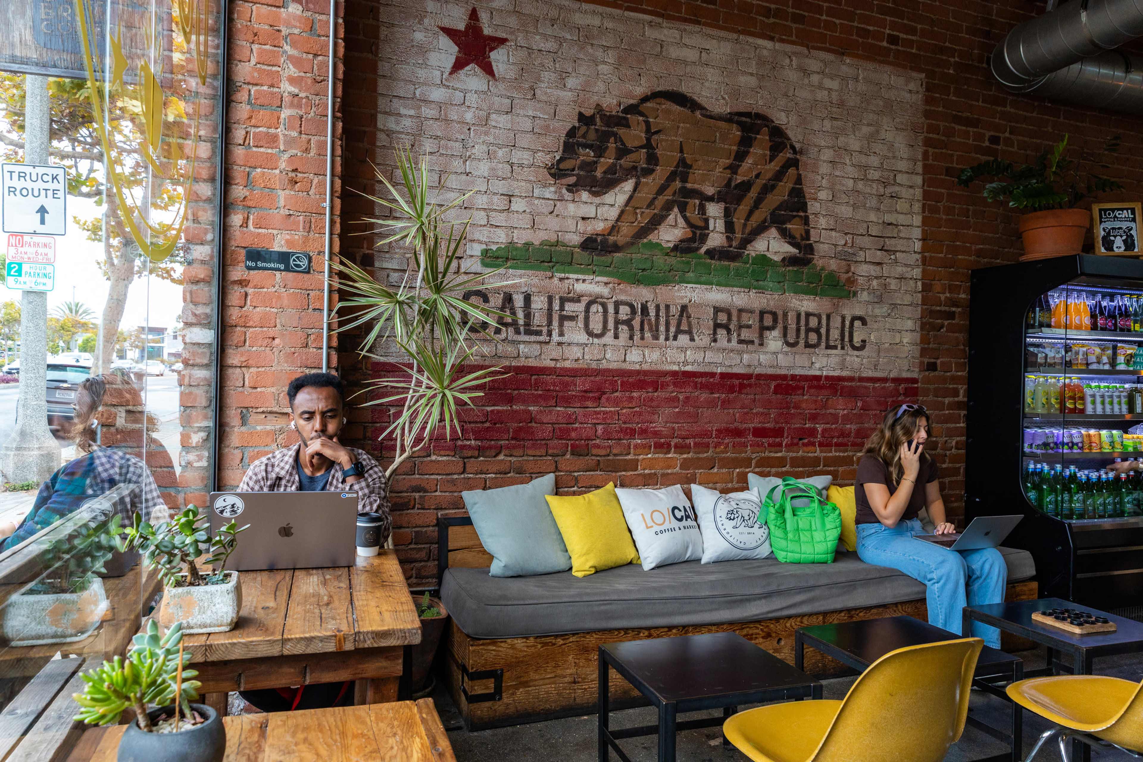 Two people work on laptops in a cafe featuring a large California Republic mural on a brick wall.