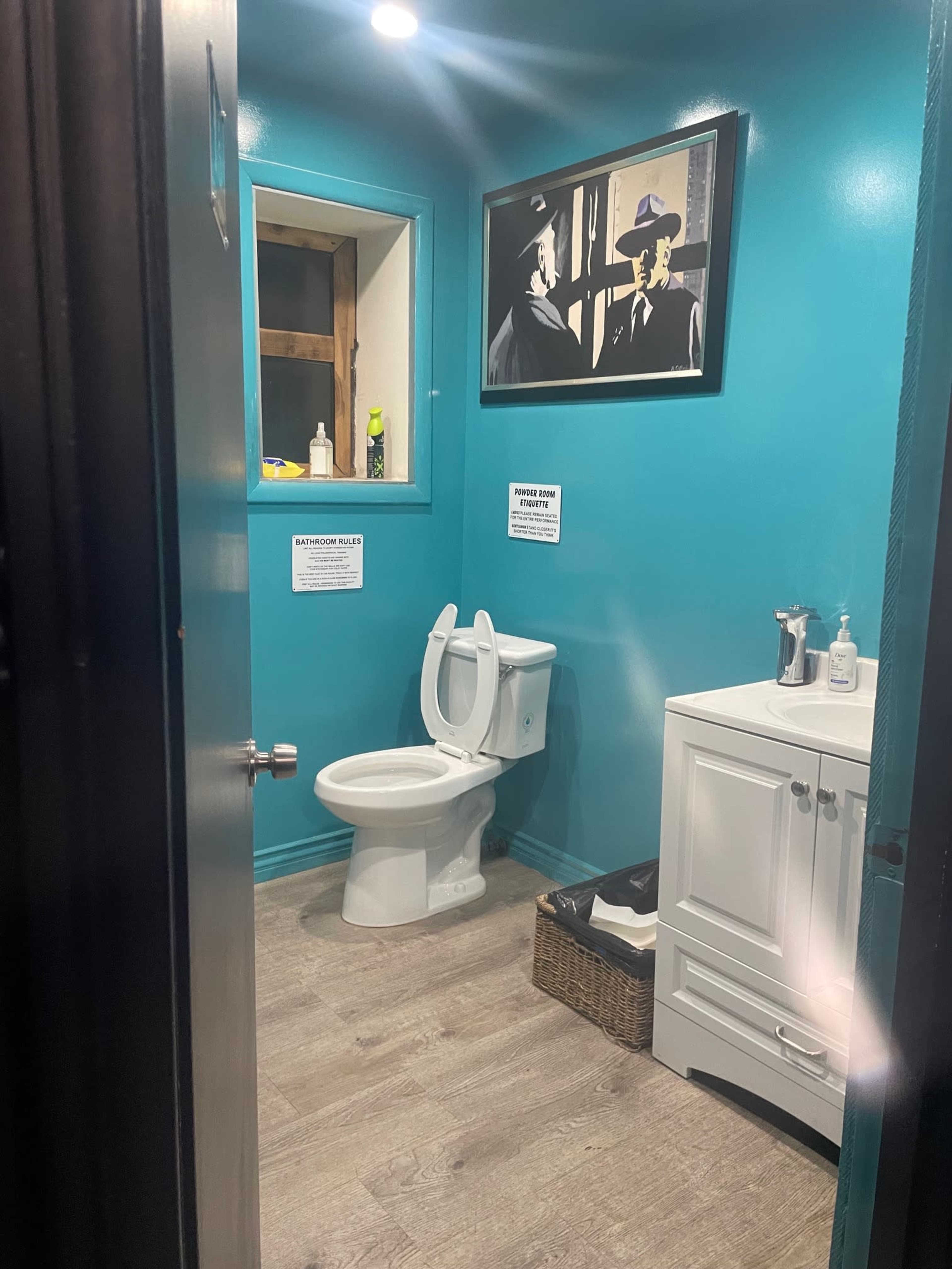 A bathroom with a turquoise wall, a white toilet, a small sink with a mirror, and a framed black-and-white photo on the wall.