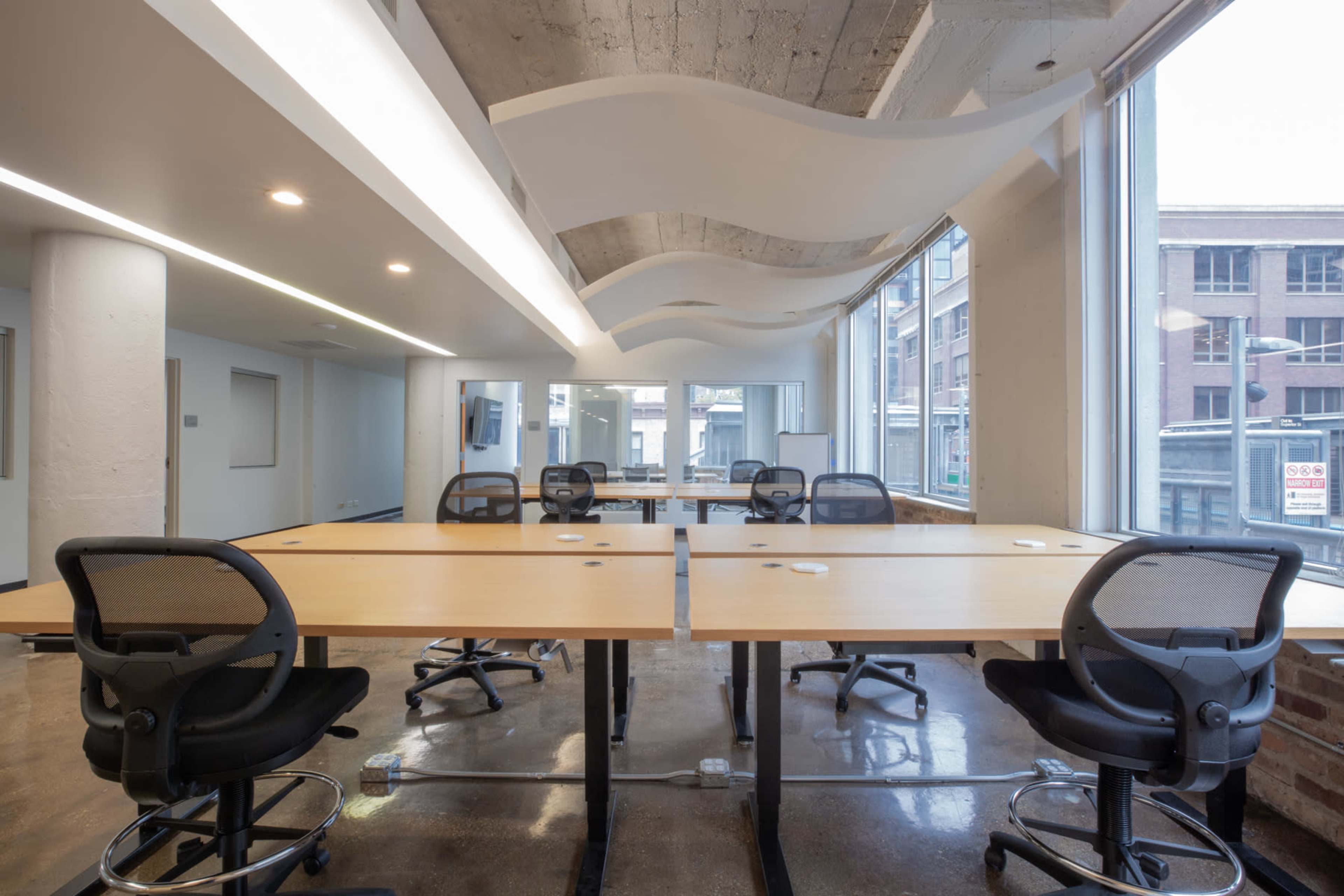 The image shows a modern office space with several tables and ergonomic chairs arranged throughout the room, featuring large windows that allow natural light to enter.