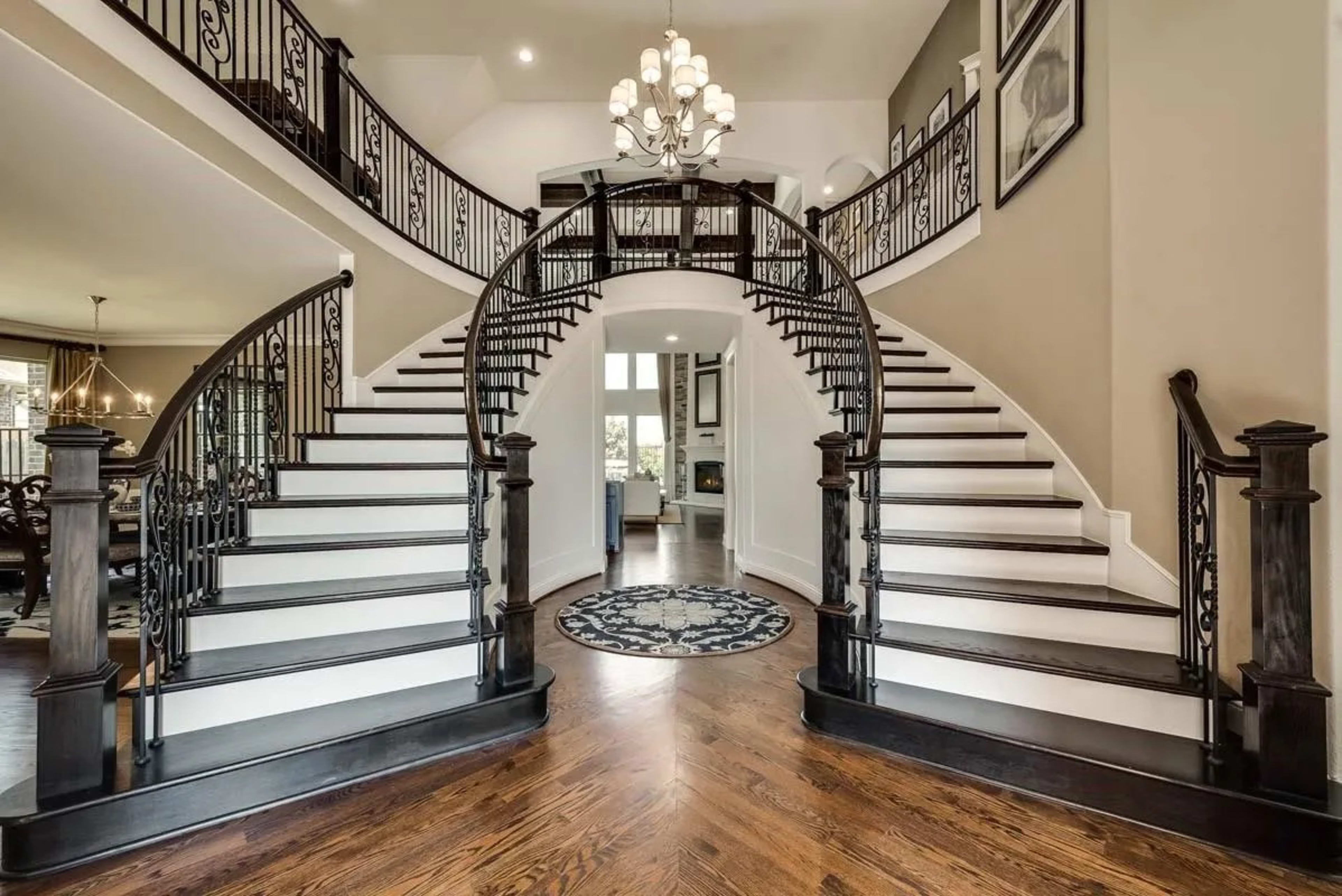 The image features a grand foyer with a double staircase, ornate railings, and a chandelier, leading into a spacious living area.